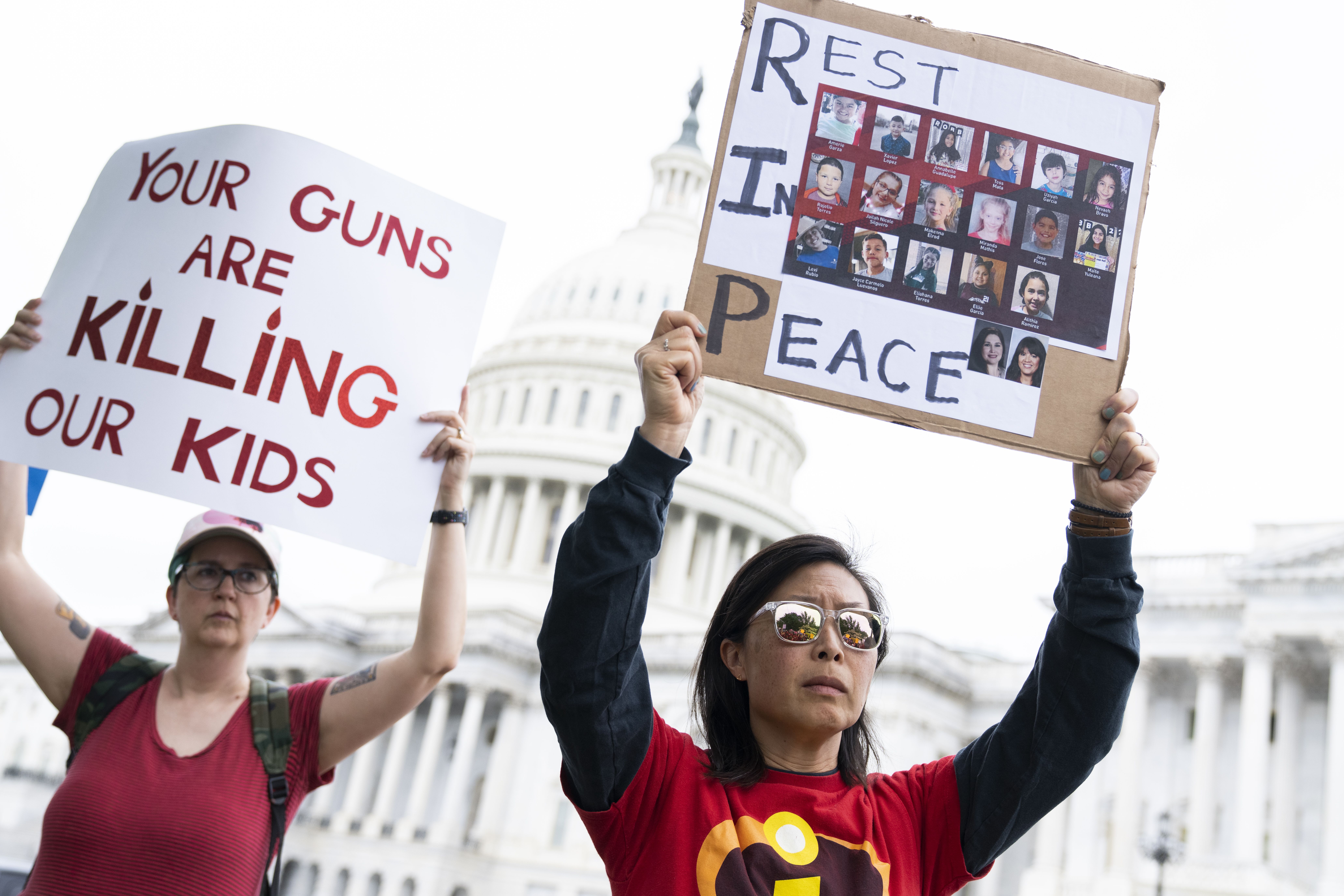 Photo of a person holding a sign with pictures of the victims of the Uvalde shooting while another person holds a sign that says "Your guns are killing our children"