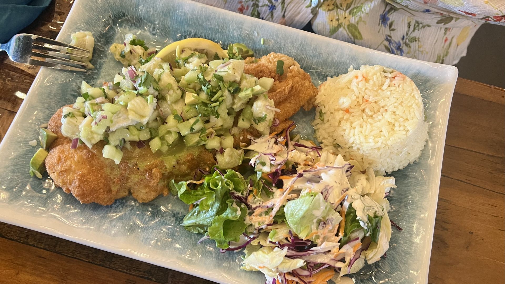 Blue rectangular plate with crispy breaded chicken topped by a chunky cucumber relish, a mound of white rice, and creamy coleslaw with shredded cabbage and greens; fork on the side.
