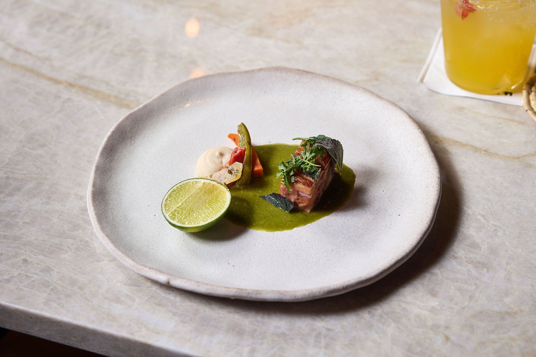 Gourmet plate with sliced meat on bright green sauce, lime wedge, herbs, and small vegetables; a yellow drink in a glass sits to the right on a marble table.