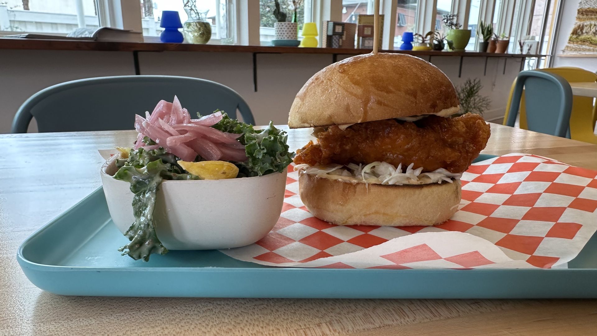 A fried chicken thigh sandwich sits on a tray with red and white checked paper. Next to the sandwich is a kale salad in a small square bowl with pickled red onions on top.