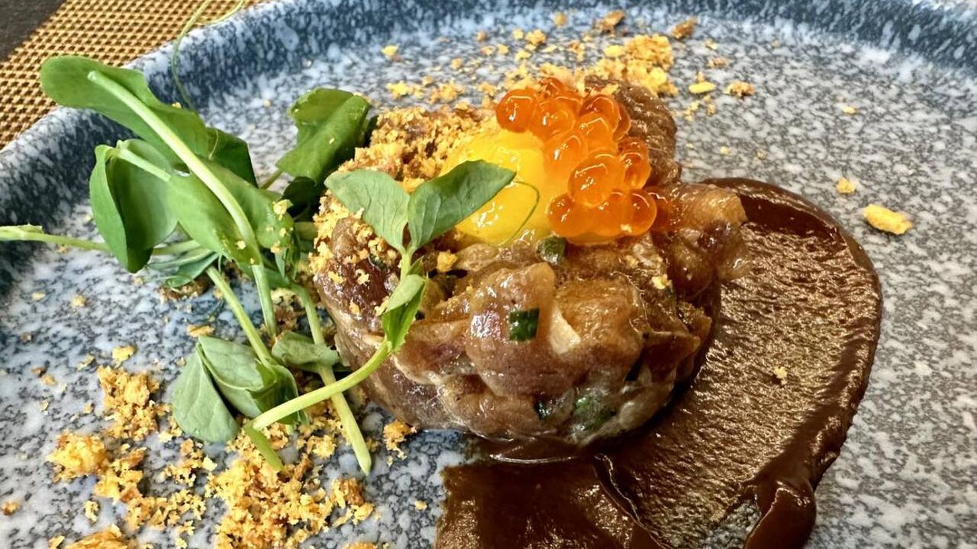 Photo shows tartare on a plate with brown sauce, egg, roe and greens.