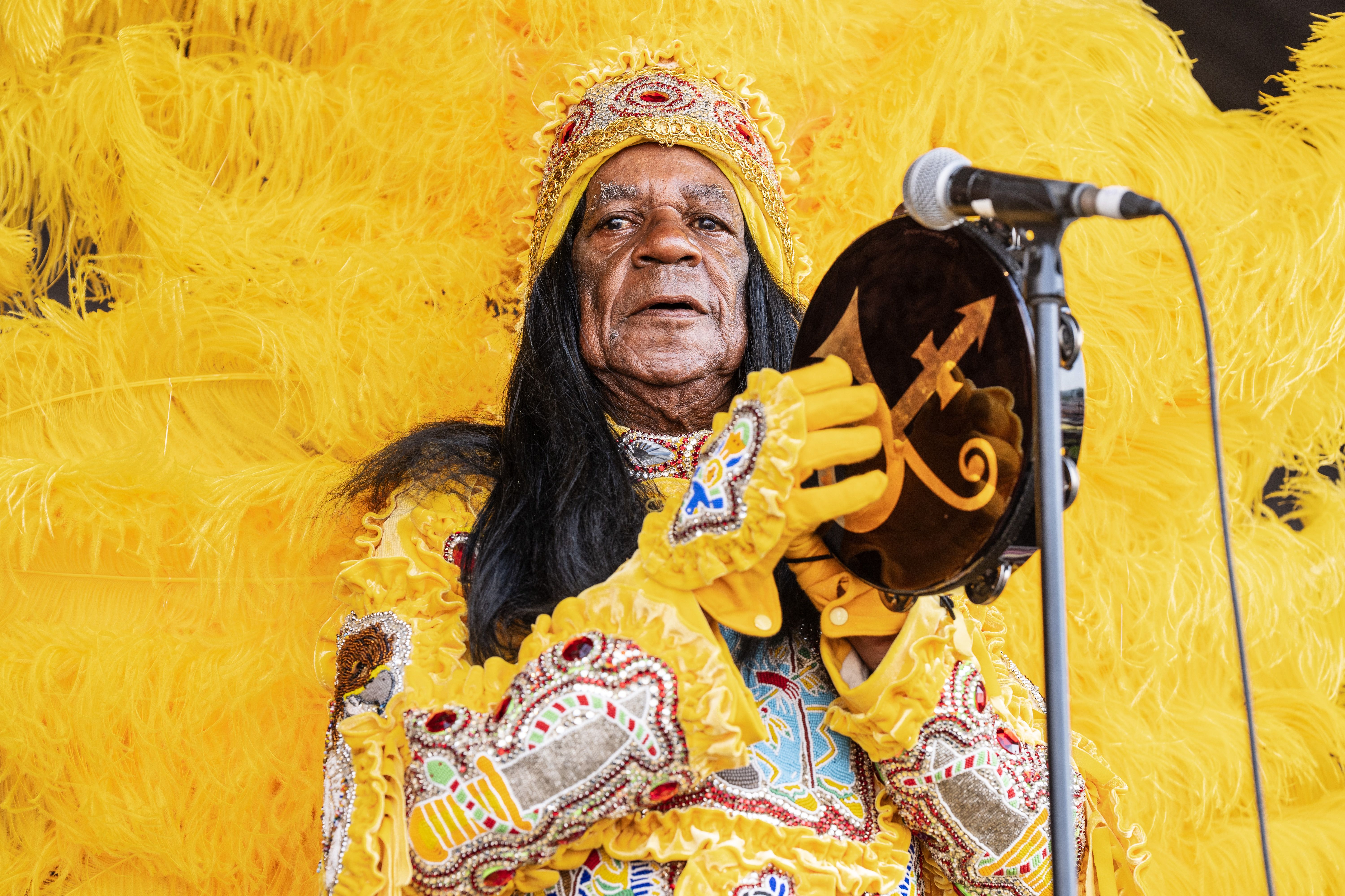 Monk Boudreaux, wearing a bright yellow-feathered Mardi Gras Indian suit, bangs a tambourine in front of a microphone.