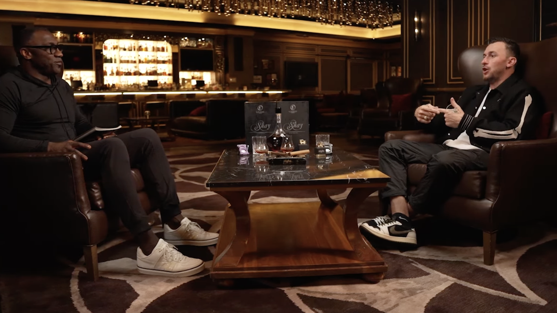 Two men sit on either side of a glass coffee table that has some booze on it