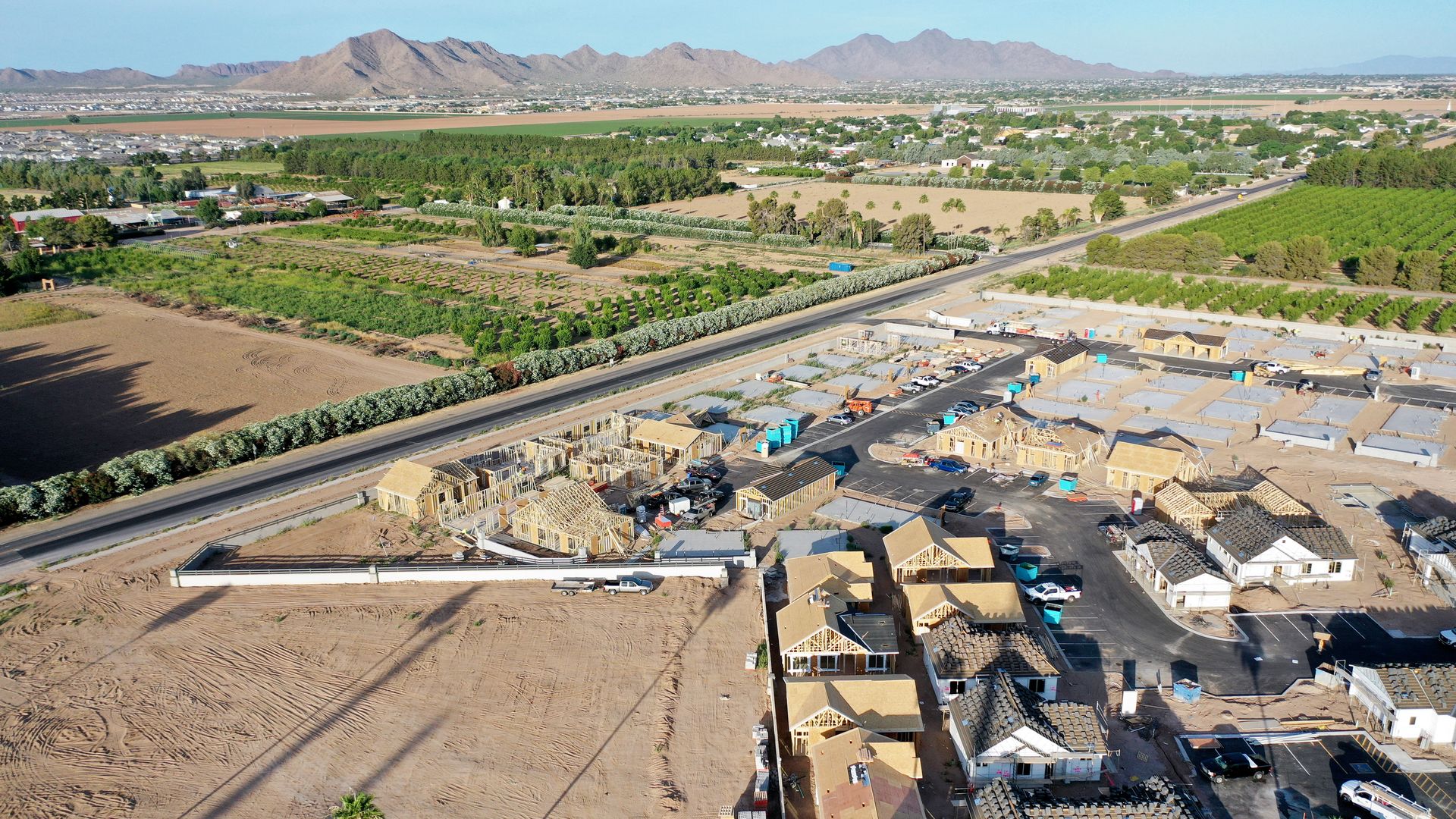 Aerial view of a partially constructed housing subdivision in a desert landscape, with trees and bushes lining undeveloped areas. Mountains and more houses are visible in the background.