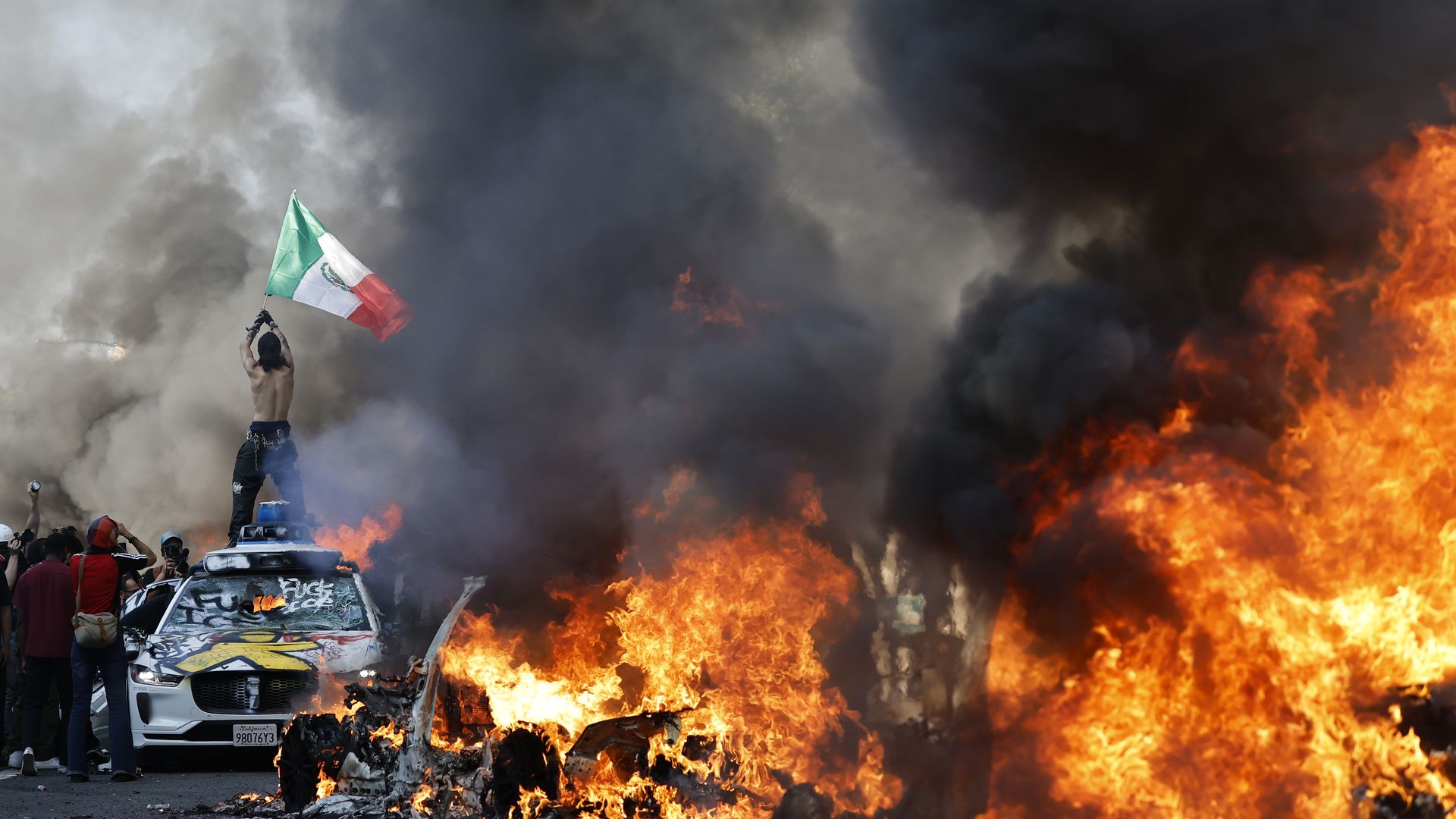 A protestor with a Mexican flag atop a burning car with burning cars in the foreground.