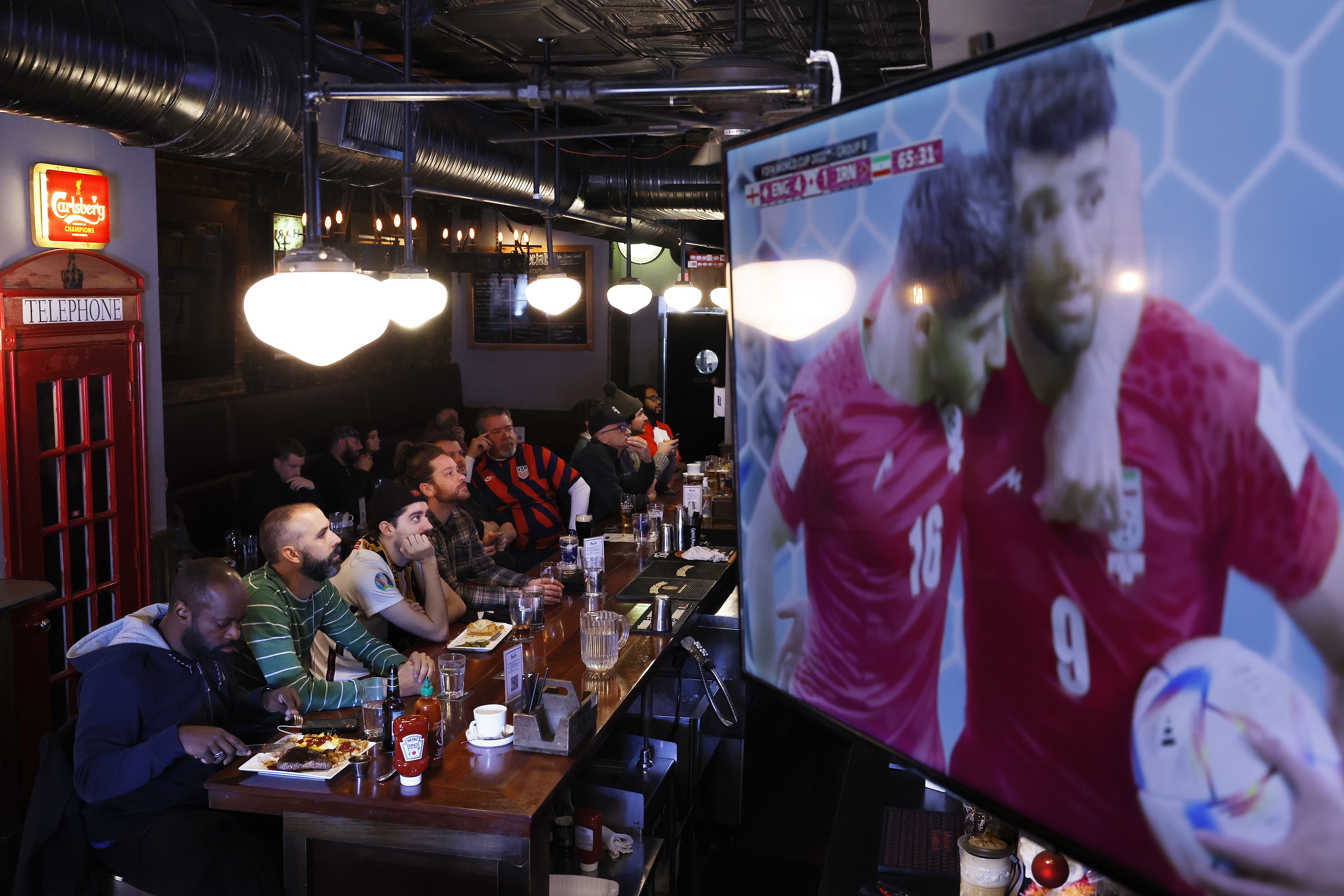 Crowded sports bar with fans at a long wooden table watching a soccer match on a large screen; a red telephone booth sign on the left, Carlsberg sign, exposed ducts, and players in pink jerseys on screen.