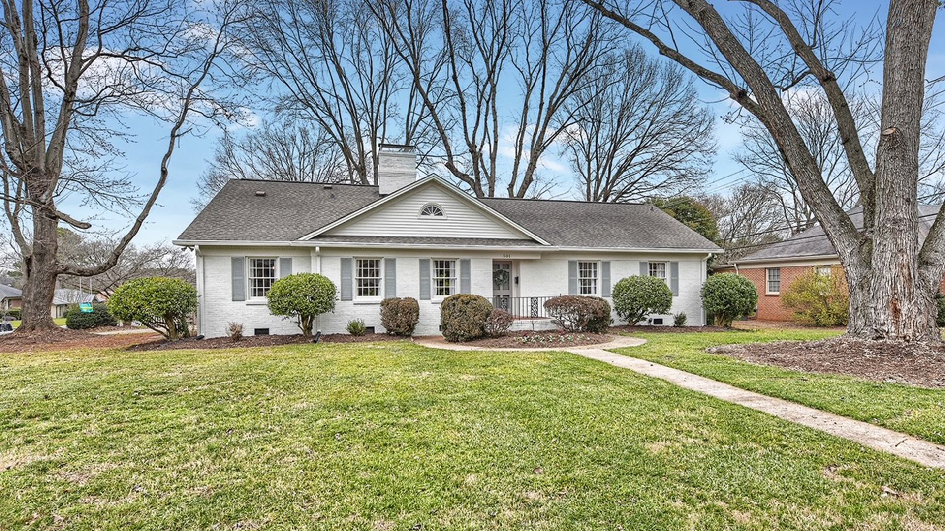 single-story home with painted-white brick and gray shutters, with a wide green yard and a walkway to the front door