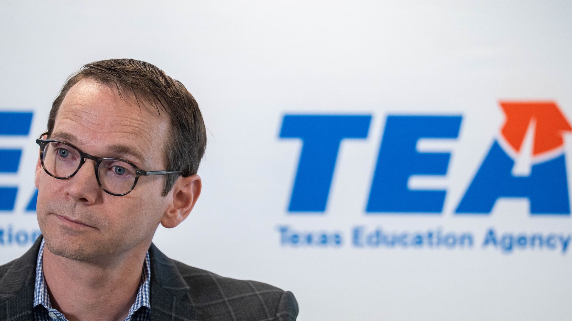 TEA Commissioner Mike Morath stands in front of a white backdrop with the blue and orange Texas Education Agency logo.