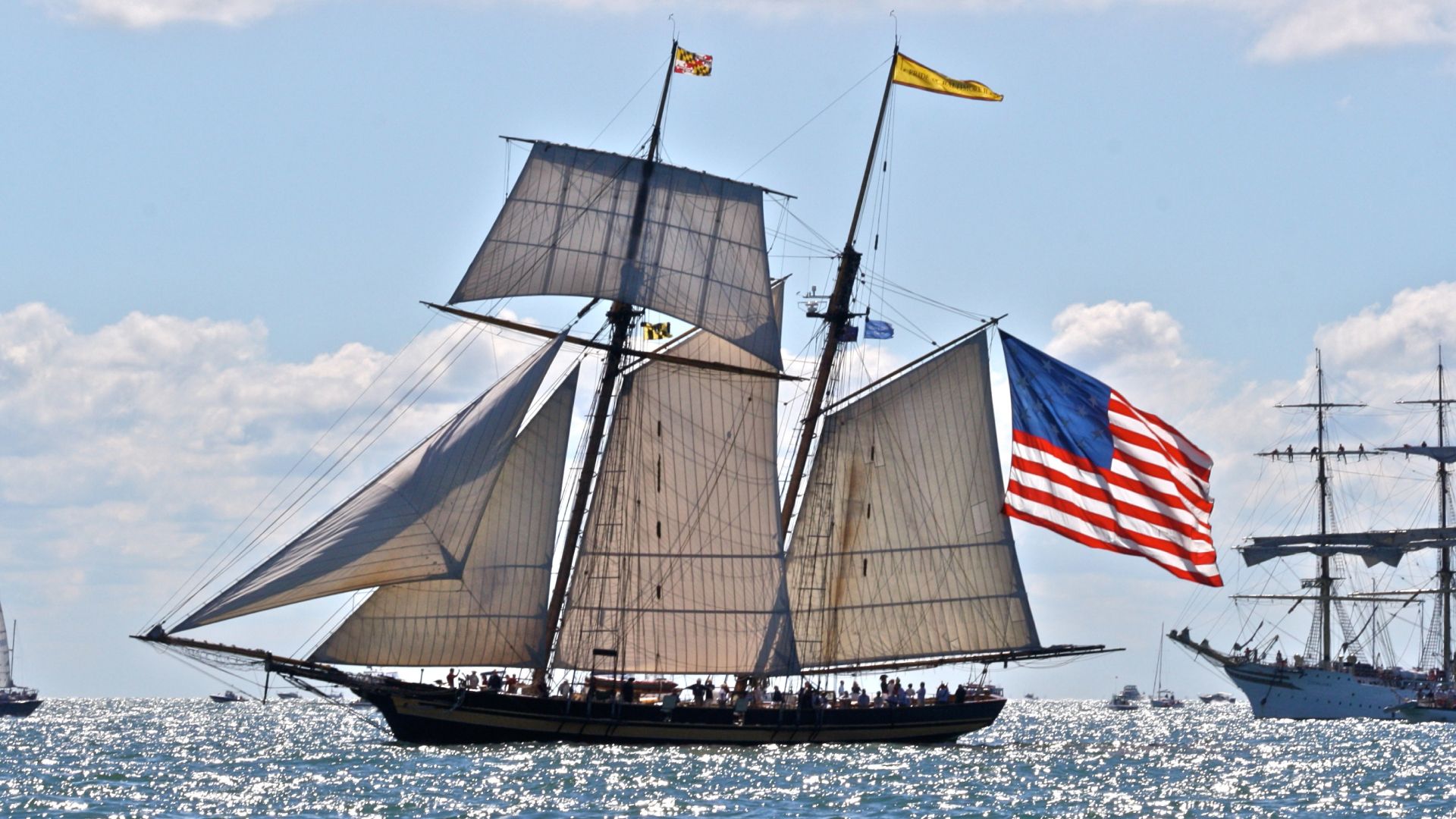 Large sailing ship with tall masts and white sails, an American flag at the stern and yellow pennants on the rigging, gliding over a sparkling blue sea under a sunny sky.