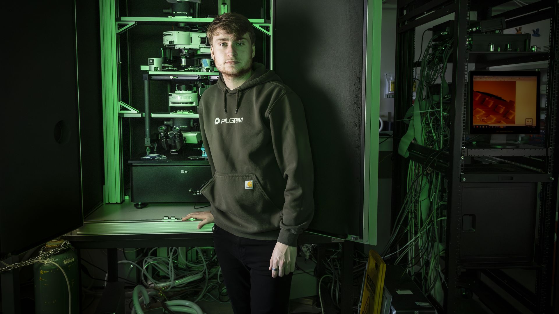 A man in a hoodie stands near machinery in a lab, lit green.
