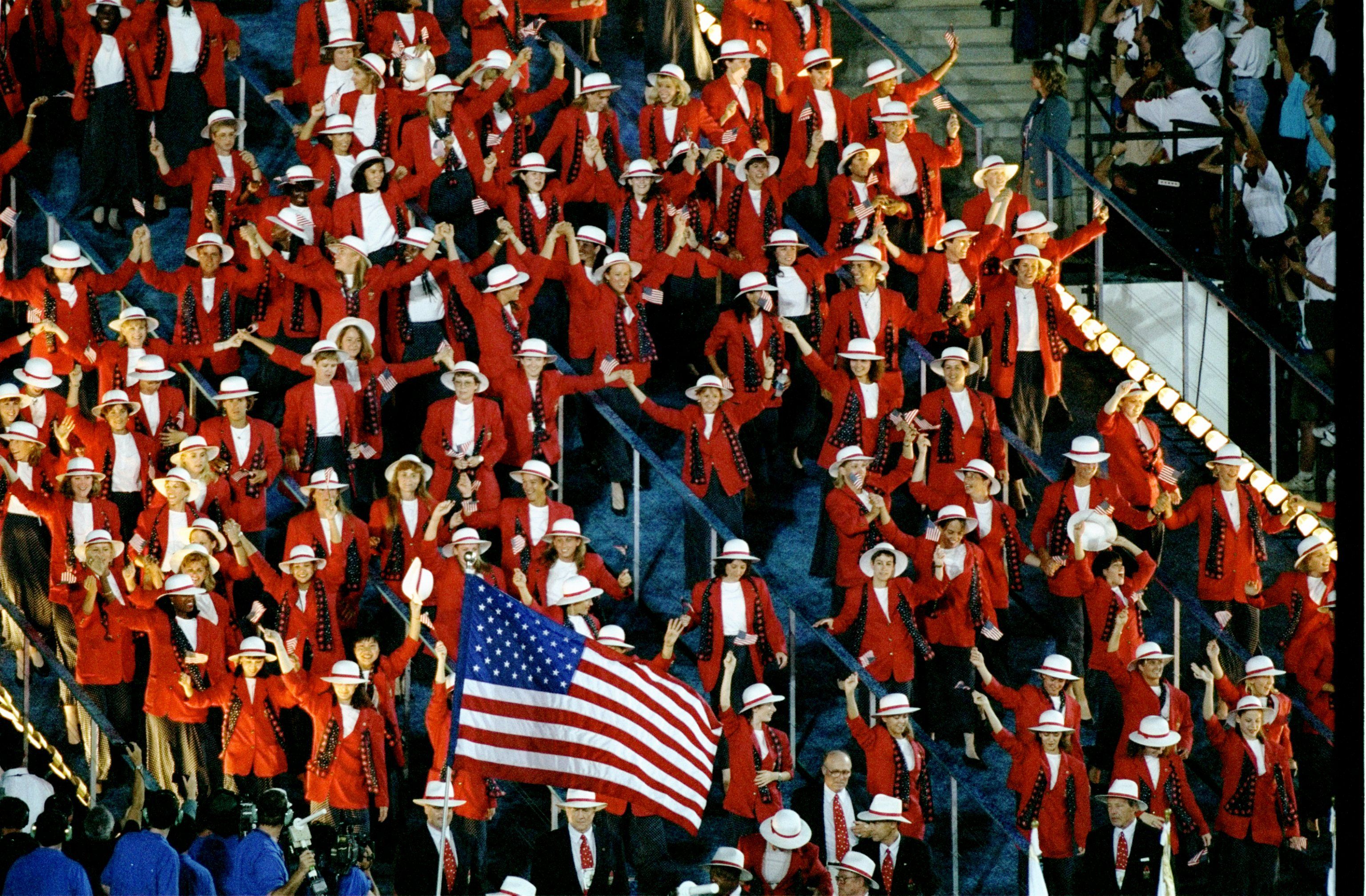 A group of dozens of athletes wearing red coats and derby hats and holding a U.S. flag march into a ceremony at a stadium
