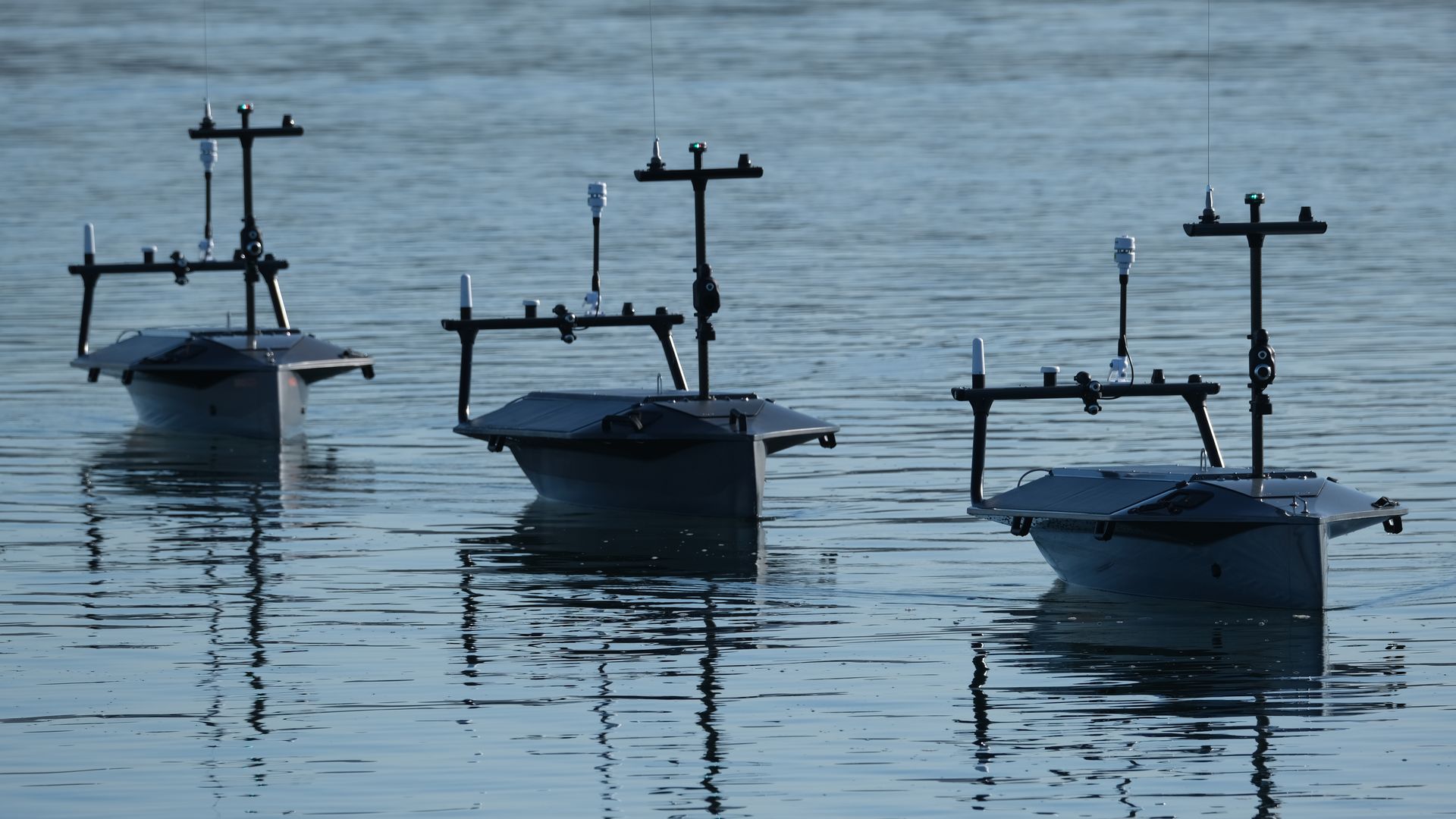Three drone boats made by Seasats move through calm waters. The boats are dark in color. The water is lighter in color.