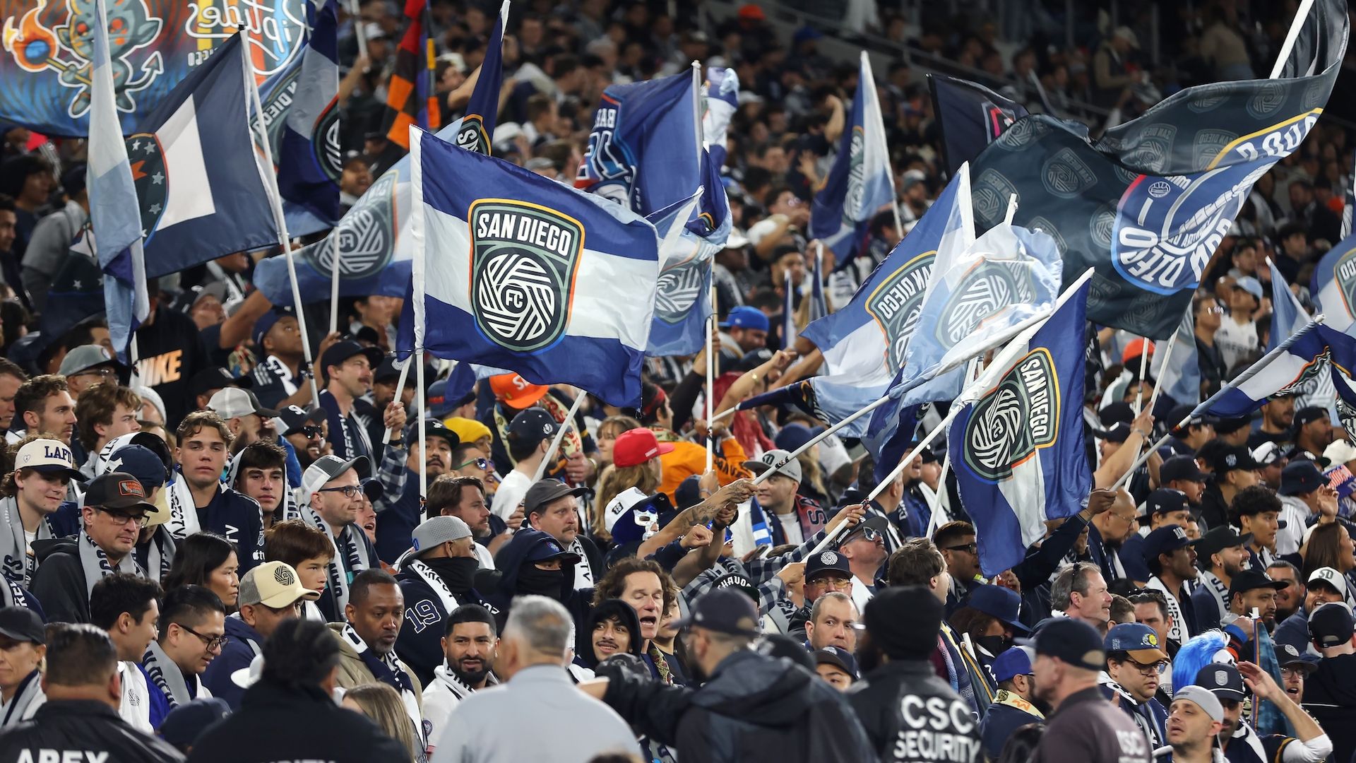 A crowded fan section with several people waving big flags at an SDFC game.