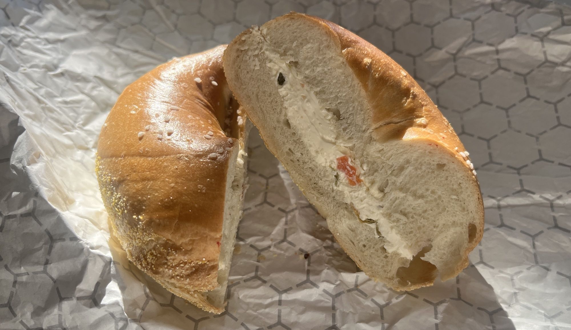 A halved sesame seed bagel with cream cheese filling, showing soft white interior and bits of red and green vegetables in the cream cheese, placed on patterned wax paper.