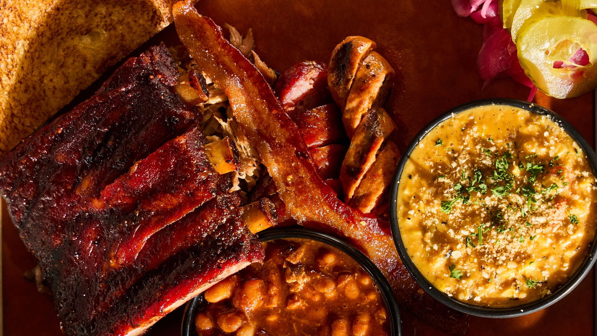 Barbecue plate with ribs, bacon strip, sliced sausage, baked beans, mac and cheese topped with herbs, pickles, and onions on a wooden tray.