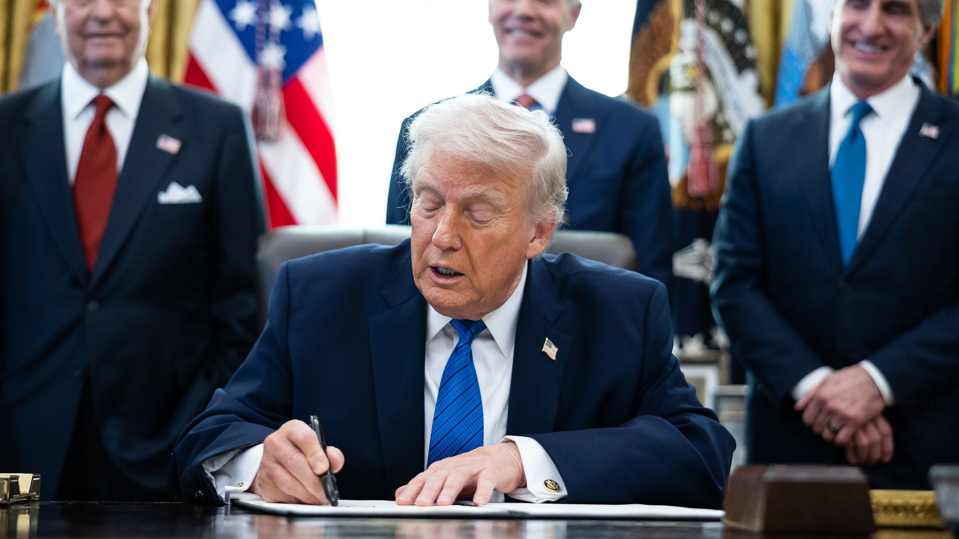 President Trump in navy suit and blue tie signs document at desk, three men in suits with red and blue ties stand behind him, American flags and presidential emblem visible.