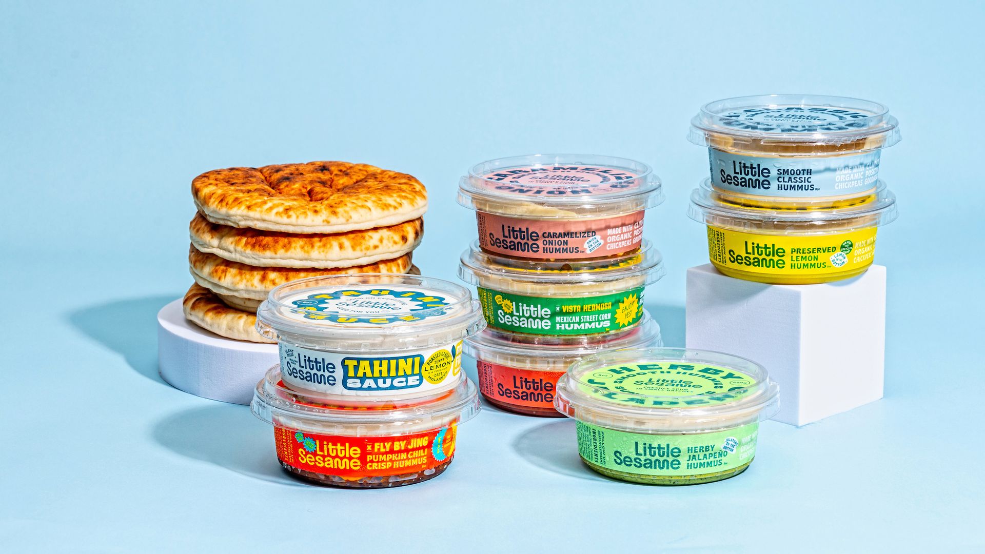 Stack of flatbreads on white pedestal beside various colorful Little Sesame hummus and tahini sauce containers on pale blue background.