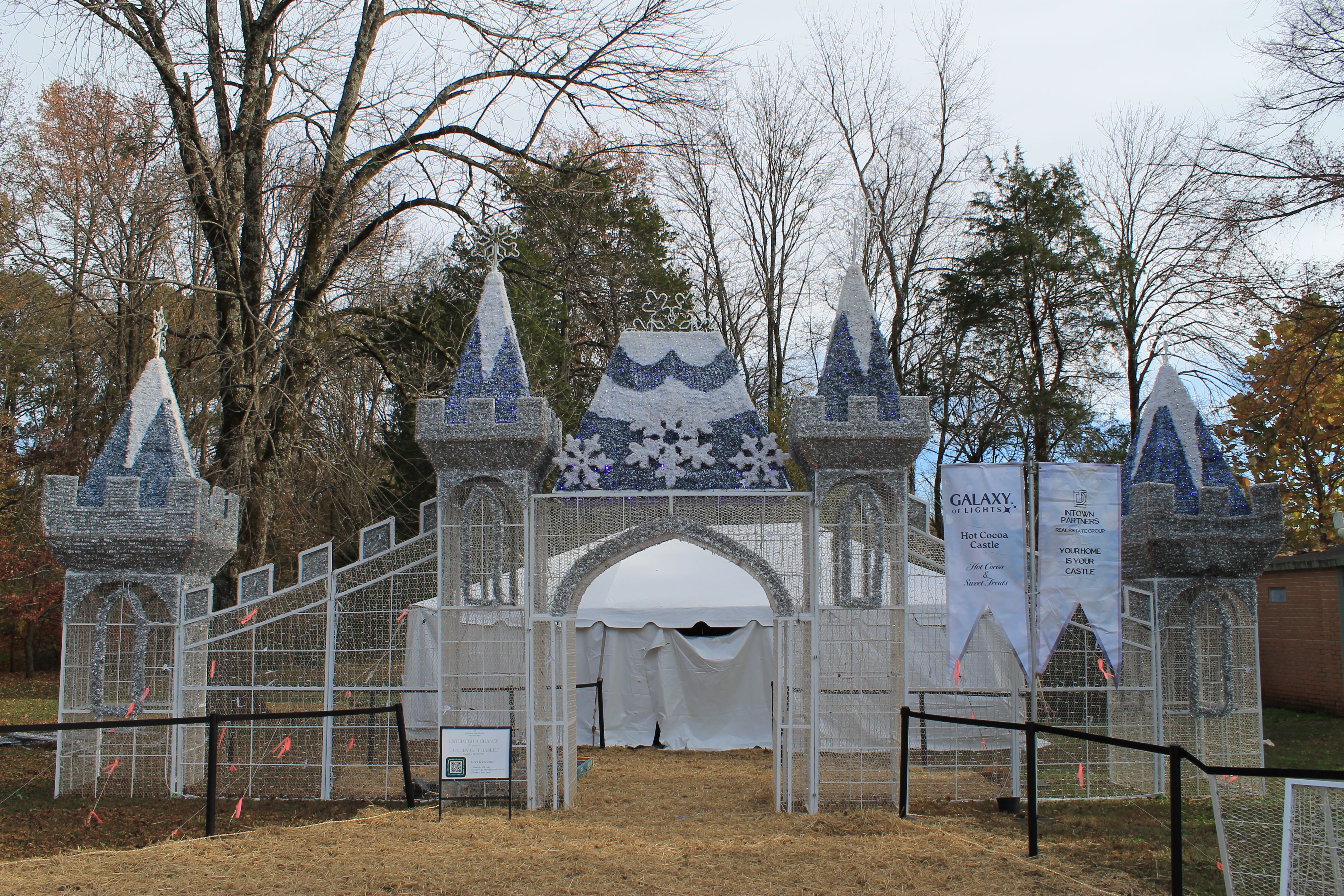 Silver and blue castle-shaped light display with snowflake decorations at an outdoor event, surrounded by bare trees and signs reading "Galaxy of Lights" and "Hot Cocoa Castle".