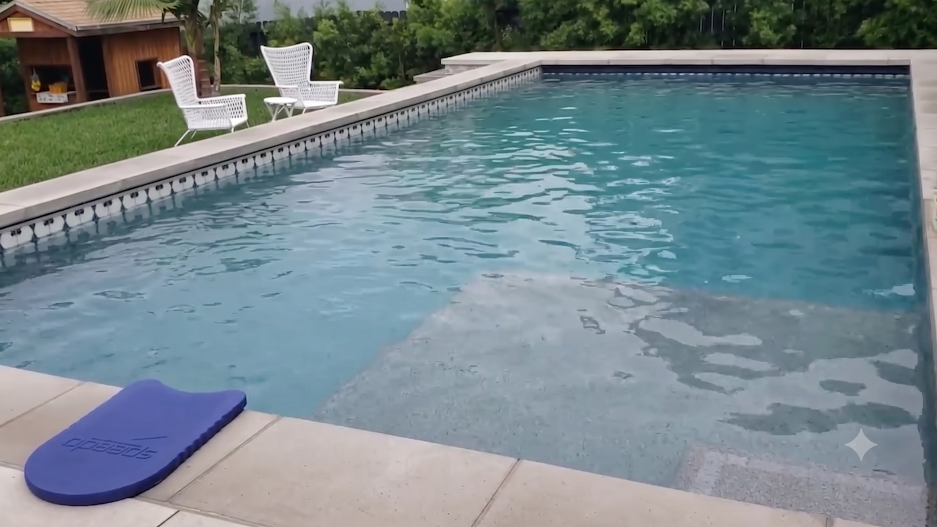 Rectangular backyard pool with clear blue water and beige deck. Two white wicker chairs and a small round table sit by the pool on a grassy lawn; a wooden shed and dense green hedges in the background.