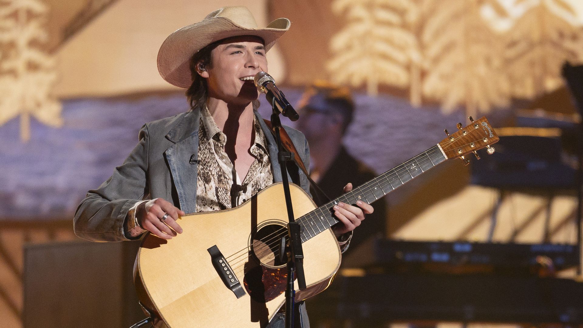 A young man sings into a microphone wearing a cowboy hat and holding an acoustic guitar.