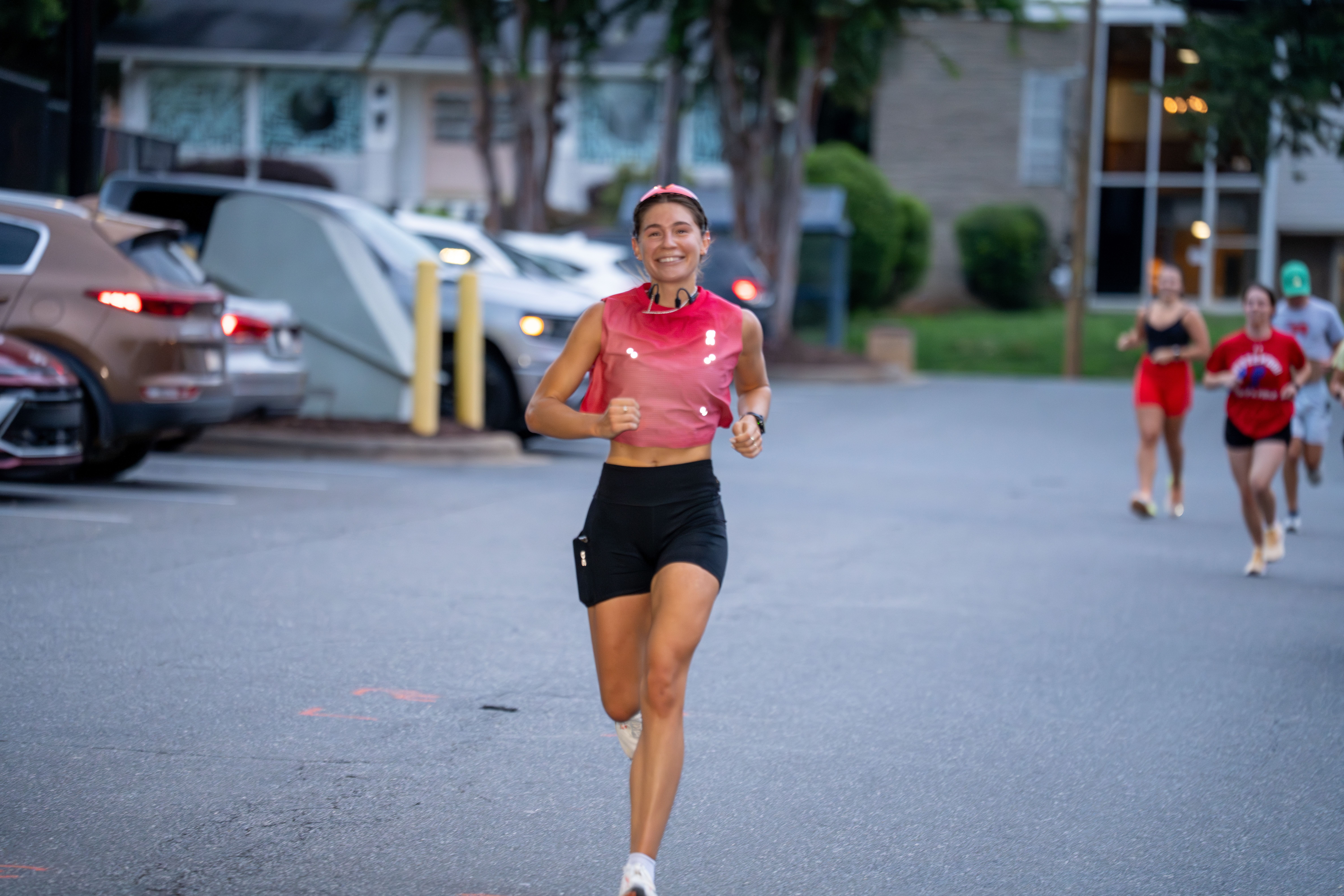 Smiling female runner in a pink sleeveless top and black shorts jogs toward the camera on a city street, with three other runners behind her and parked cars and buildings in view.