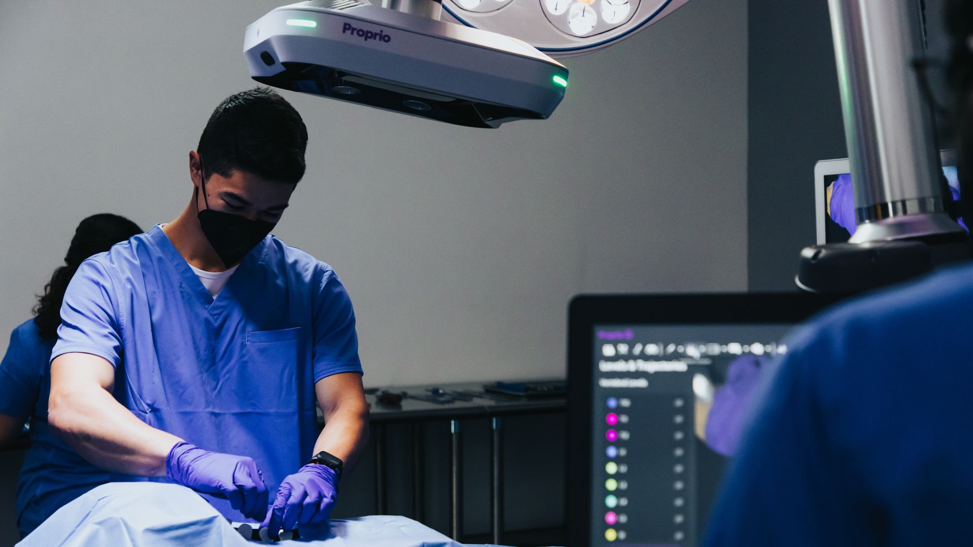 A person in a surgical mask and scrubs operates on a patient, with a device overhead and another person at a monitor nearby.