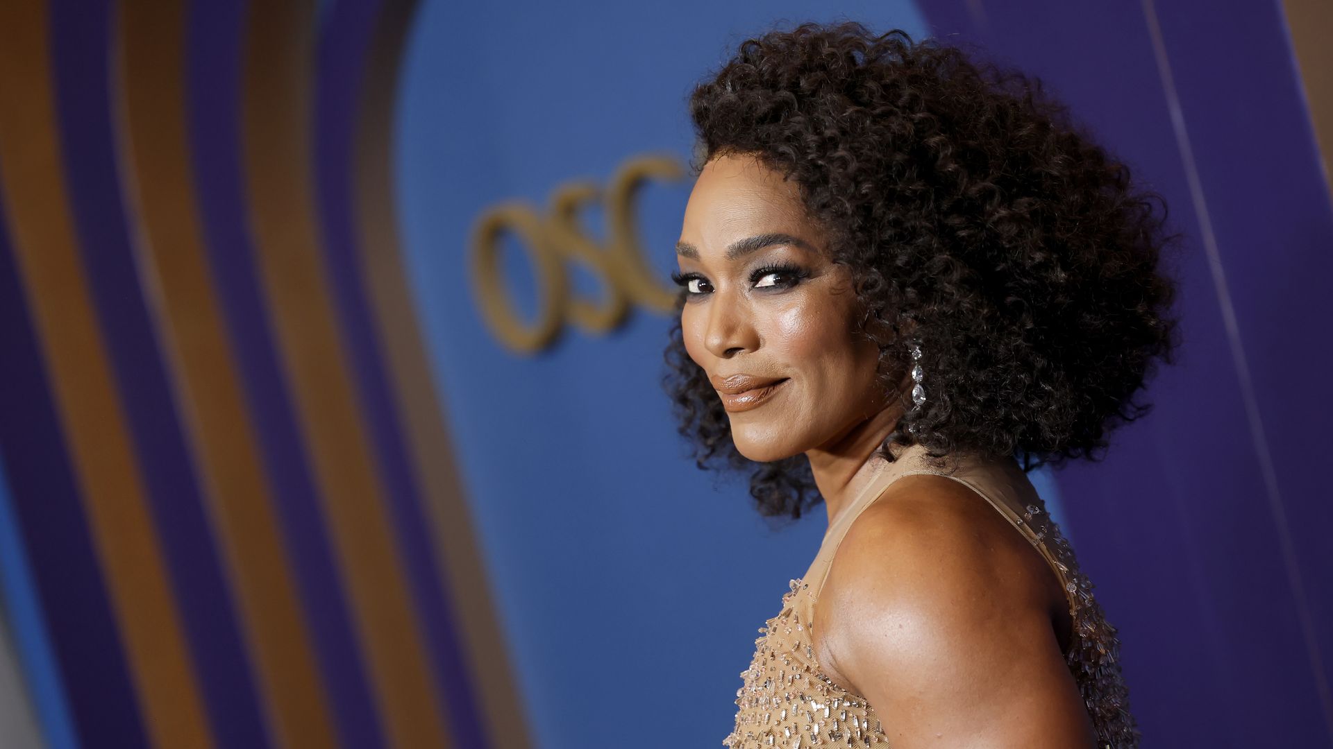Woman with curly dark hair wearing a sleeveless beige dress with sequins, posing and smiling sideways against a blue and purple backdrop with gold lettering.