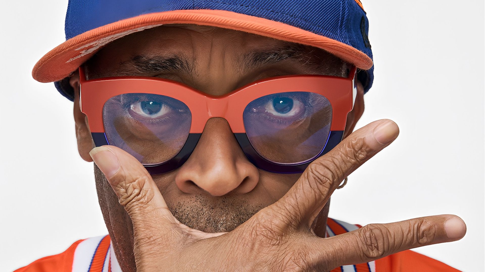 Close-up of a person wearing a blue Knicks cap with an orange basketball logo and a heart patch, oversized orange sunglasses, an orange jacket, and gold chains; NYC Nike logo in the top-right.