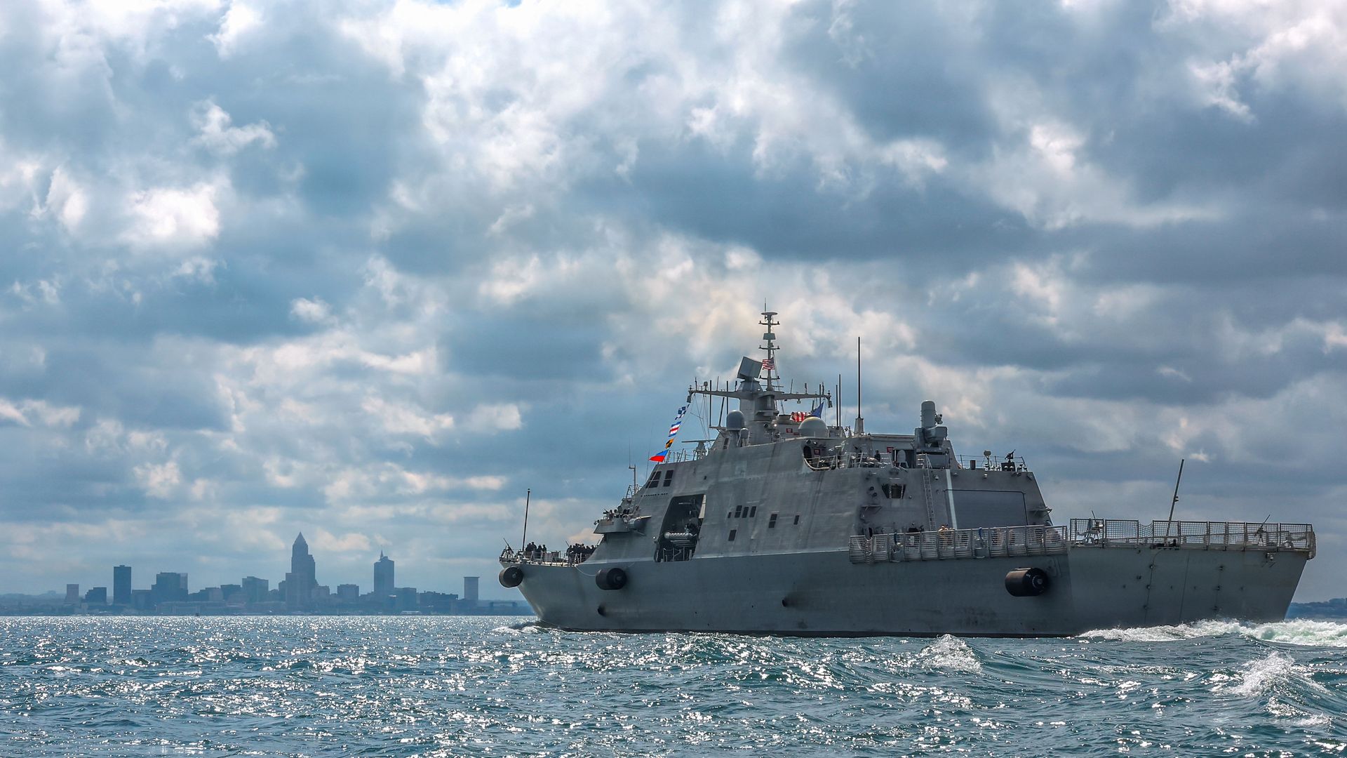 Large gray military warship glides through blue water beneath a cloudy sky, with a distant city skyline on the horizon and people visible on the deck.