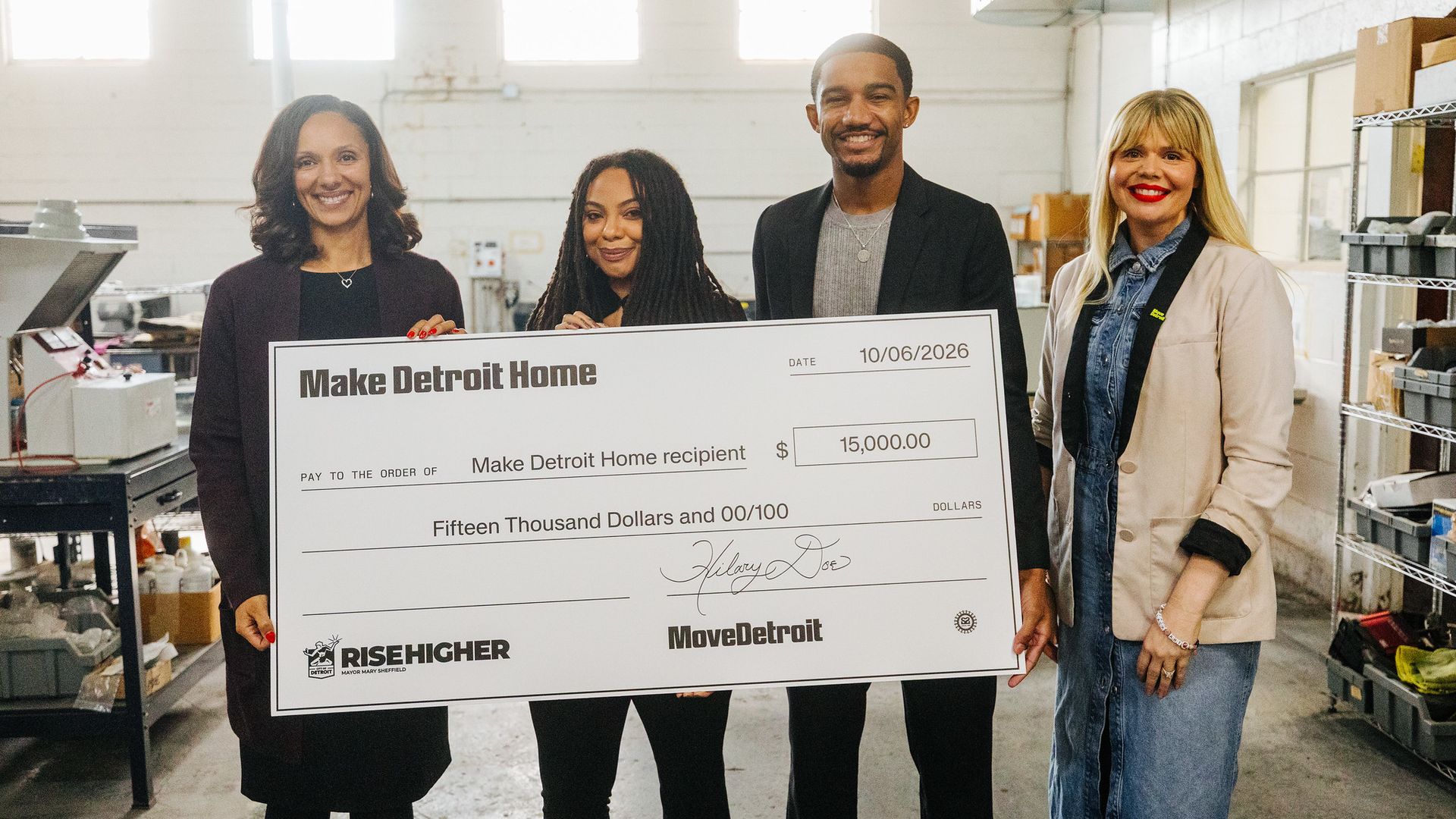 Officials and grant recipients smile in a bright warehouse, holding a large check that reads "Make Detroit Home" for $15,000; dated 10/06/2026, with shelves and equipment visible in the background.
