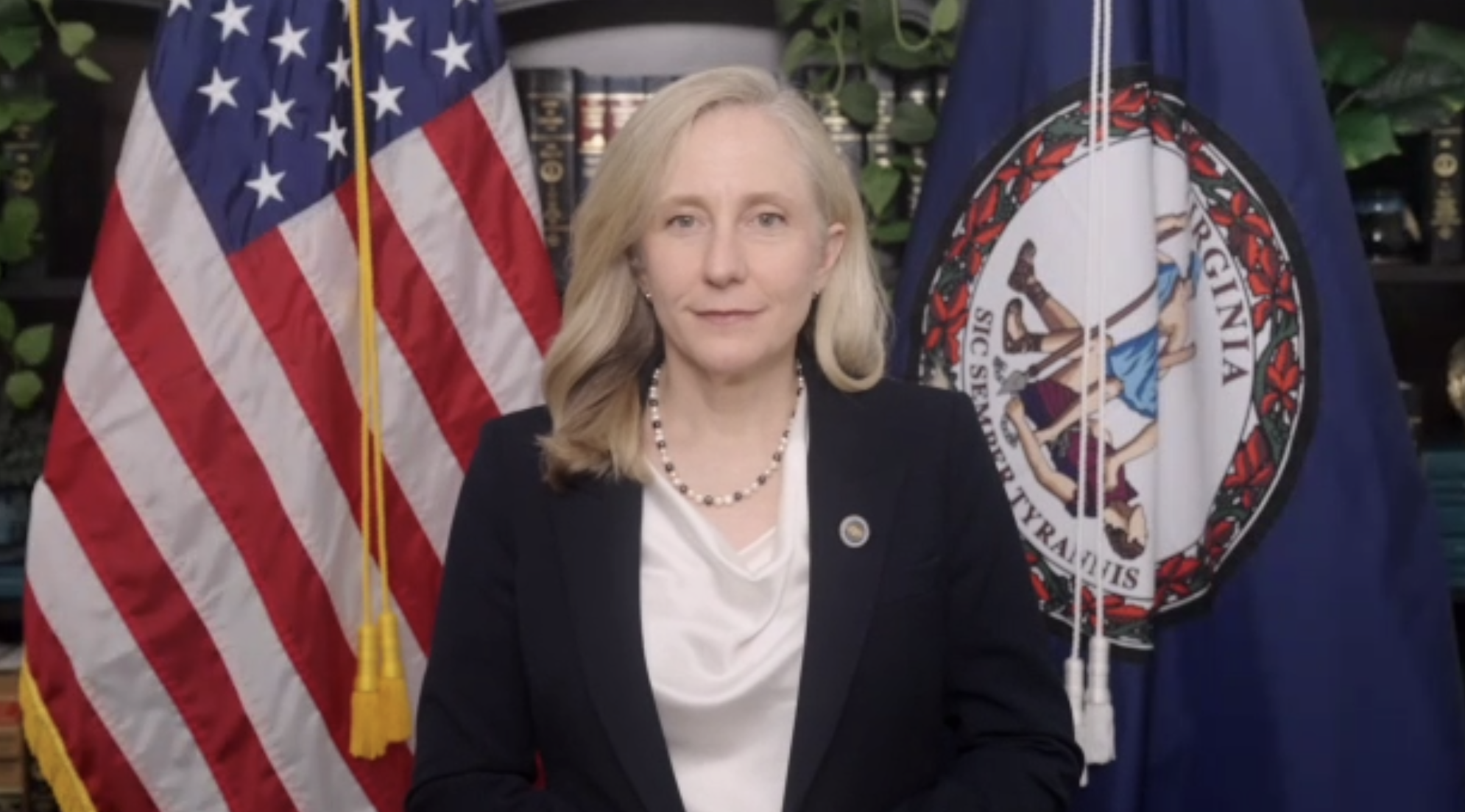 Blonde woman in a dark blazer and white blouse, wearing a pearl necklace and pin, stands in front of the U.S. flag and the Virginia state flag, with books and greenery in the background.