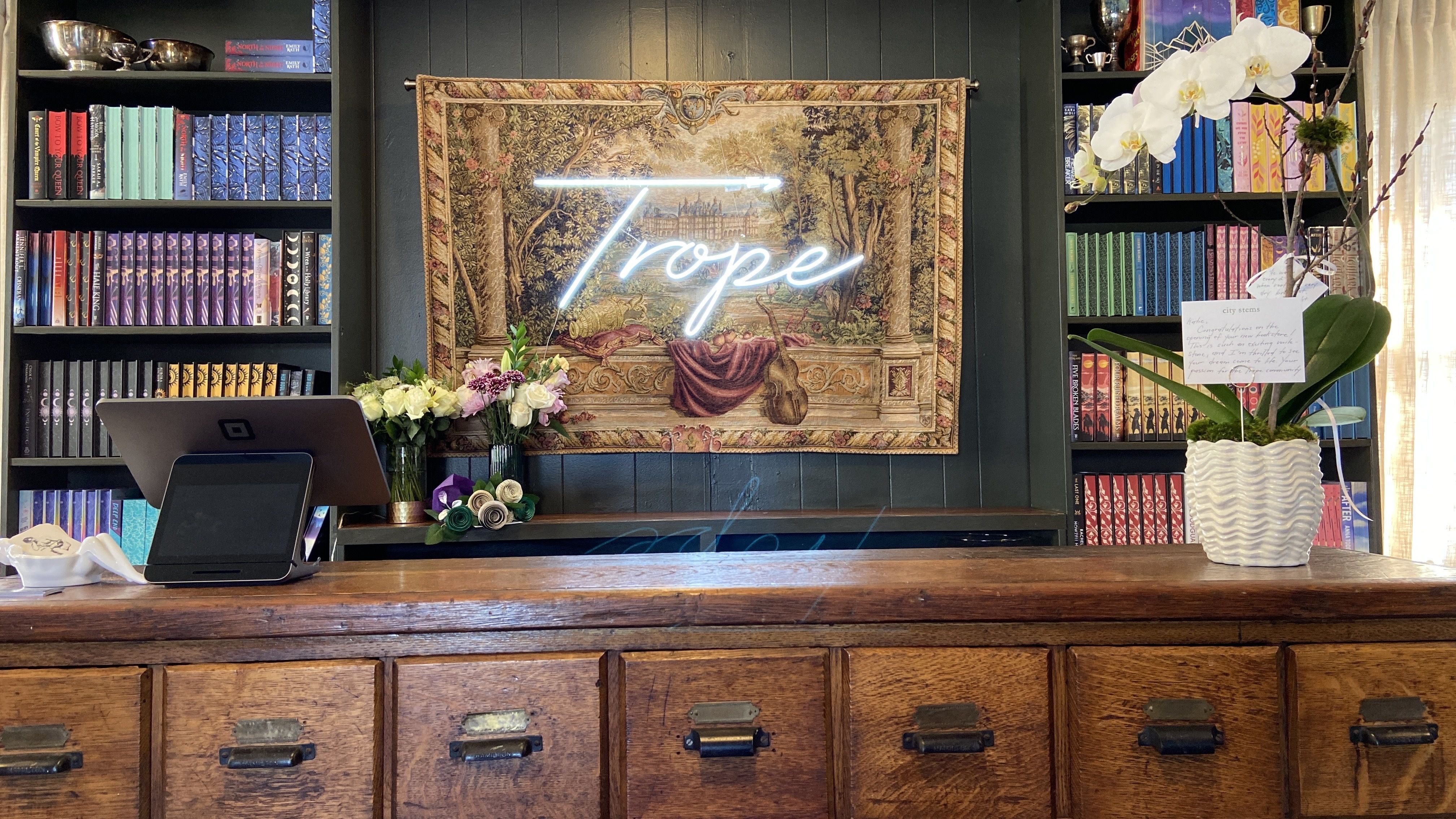 Trope Bookshop's checkout counter is an antique piece with a tapestry with the word "Trope" lit up behind it. 