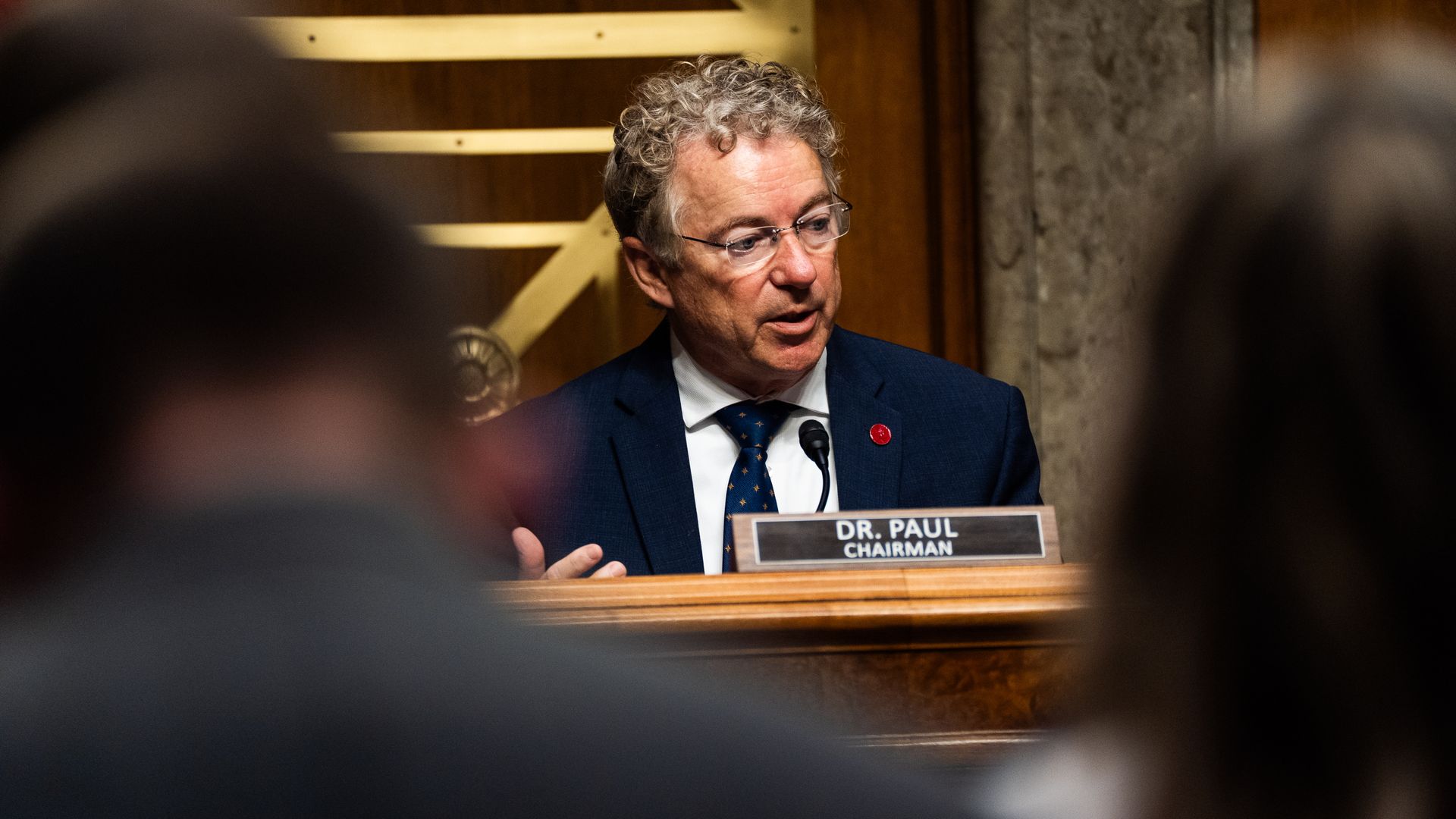 Rand Paul speaks into a microphone during a hearing on Capitol Hill. A plaque in front of him reads "Dr. Paul Chairman."