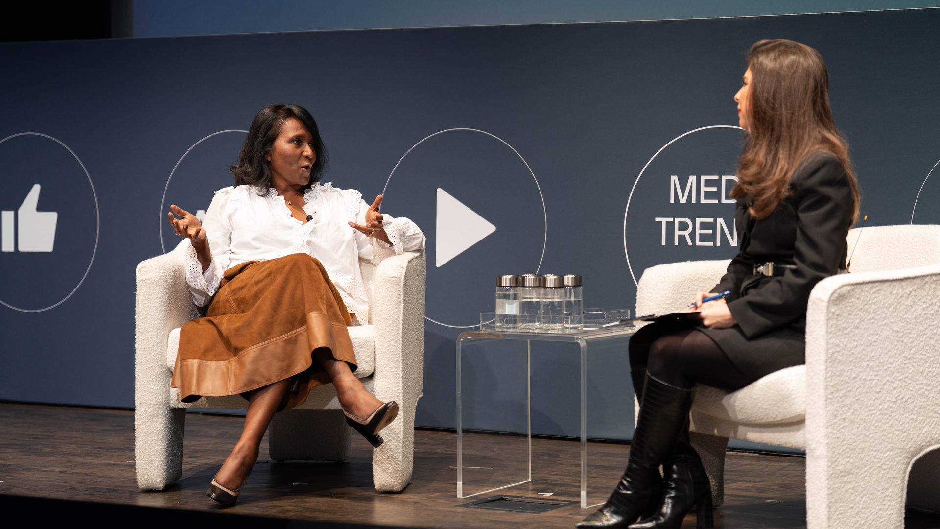 Two women seated on white armchairs on stage in conversation; left woman in white blouse and brown skirt, right woman in black jacket and boots, with a clear table holding water bottles between them.