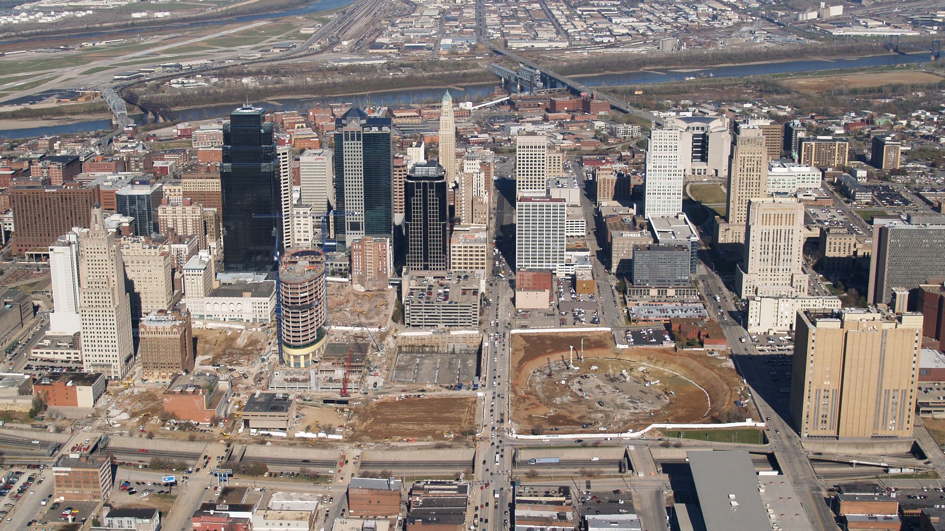 Aerial view of a city skyline with tall buildings, construction sites, roads, bridges, and a river in the background under a clear sky.