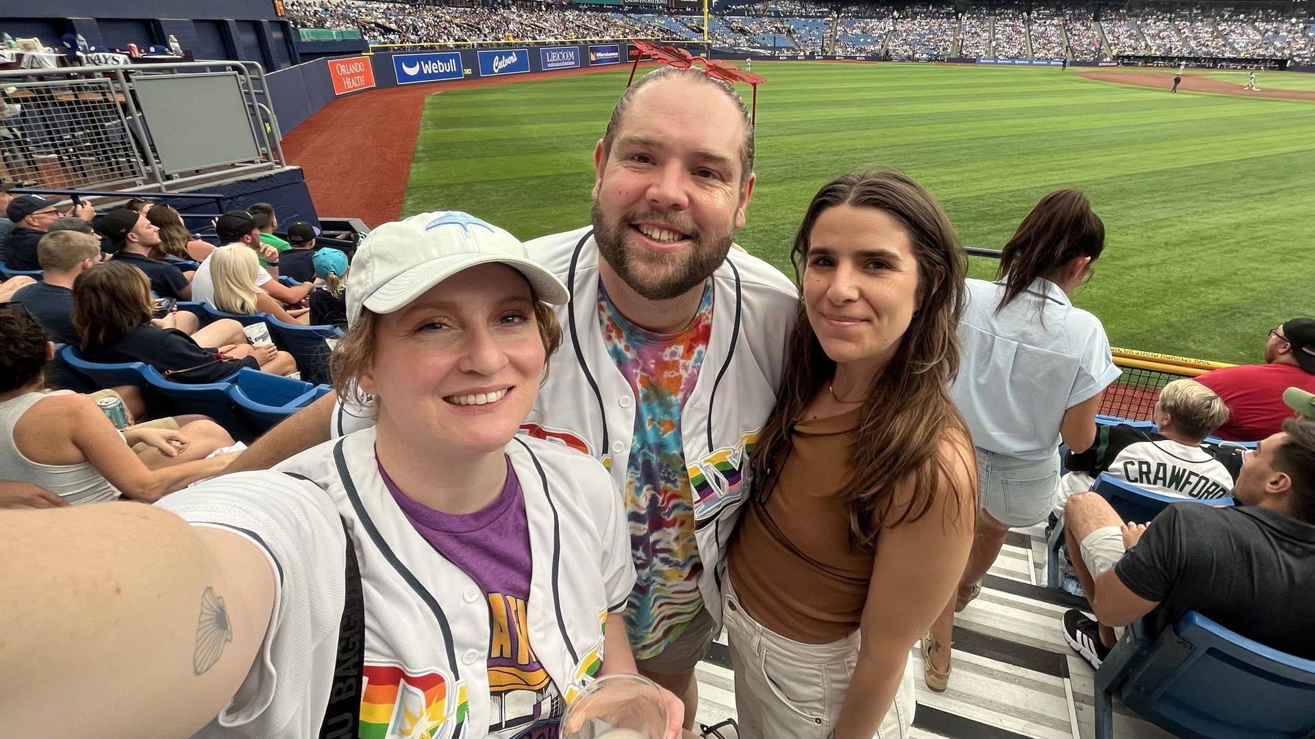 Three friends take a selfie at a baseball game: a woman in a white jersey, a woman in a brown tank top and a man in a tie-dye shirt, with the green field and crowded stands behind them.