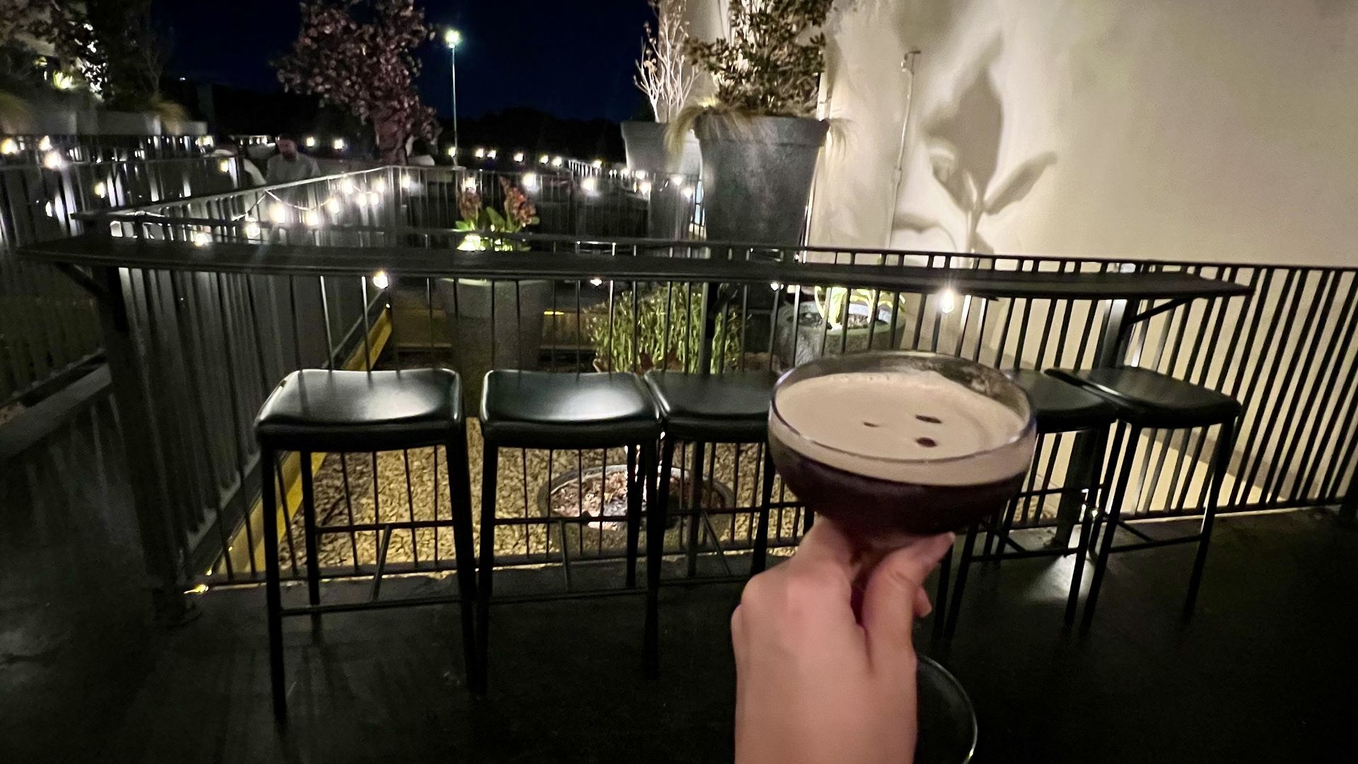 Hand holding a dark cocktail glass with foam, outdoor patio with black stools, string lights, plants, and nighttime lighting.