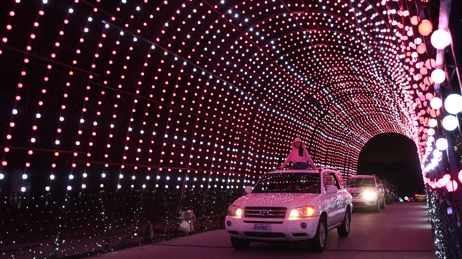 A person wearing a white hoodie sits on a white SUV's roof as it drives through a tunnel of red and white glowing light bulbs at night, with other cars following behind.