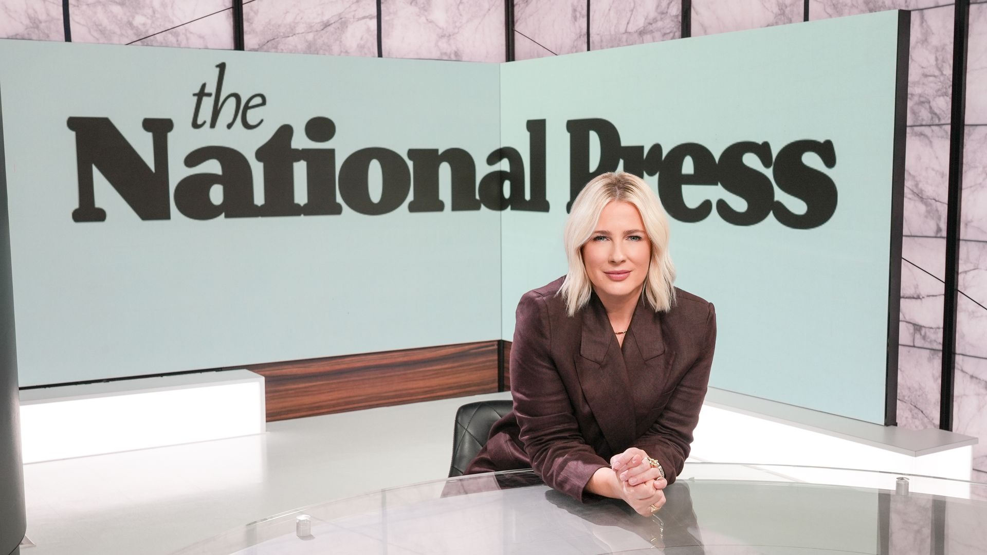 A news studio with a large sign reading "The National Press" in black on pale blue panels; a blonde woman in a burgundy blazer sits at a glass desk.