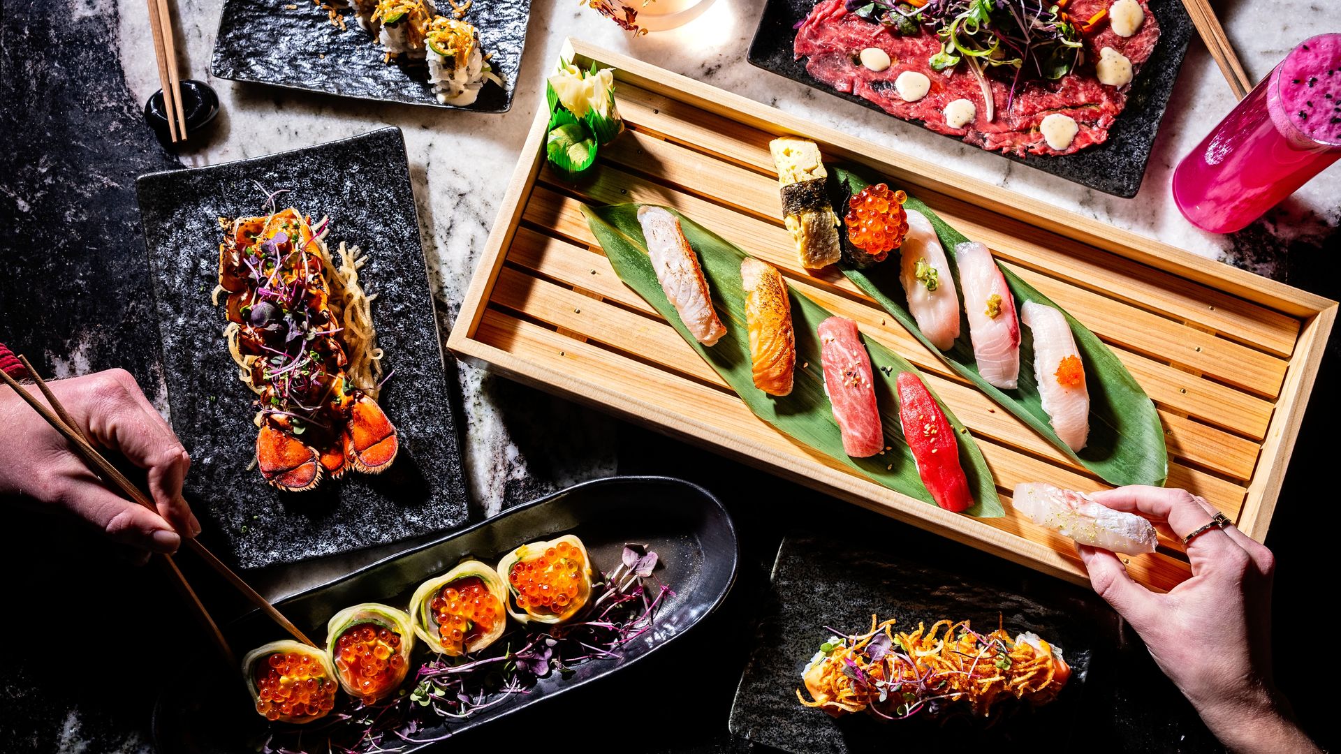 Top view of assorted sushi and sashimi on black and wooden plates with garnishes, two hands using chopsticks and holding sushi, and a bright pink drink on a marble surface.