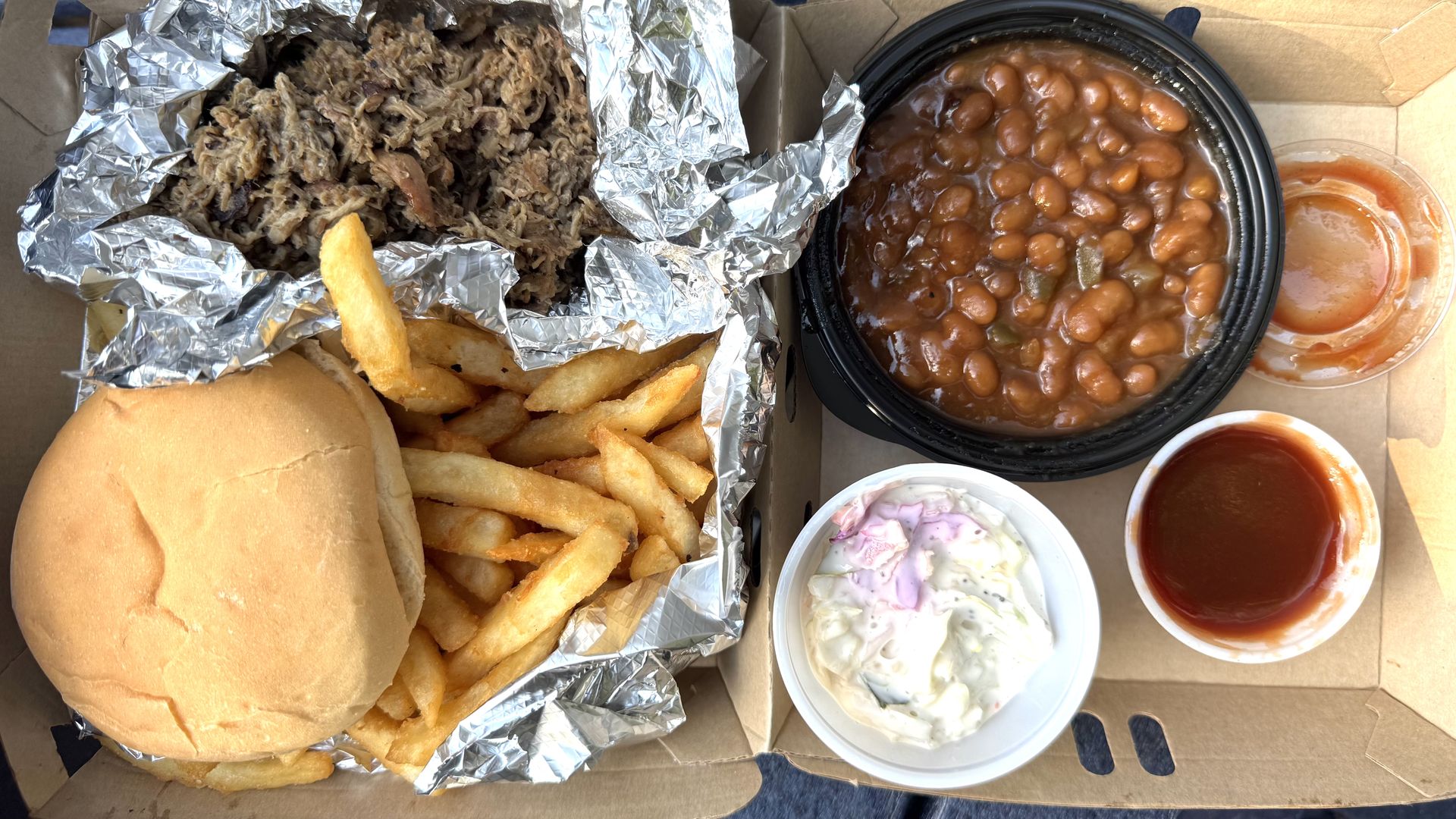 A takeout barbecue meal includes a pulled pork sandwich with fries, baked beans, coleslaw, and barbecue sauce.