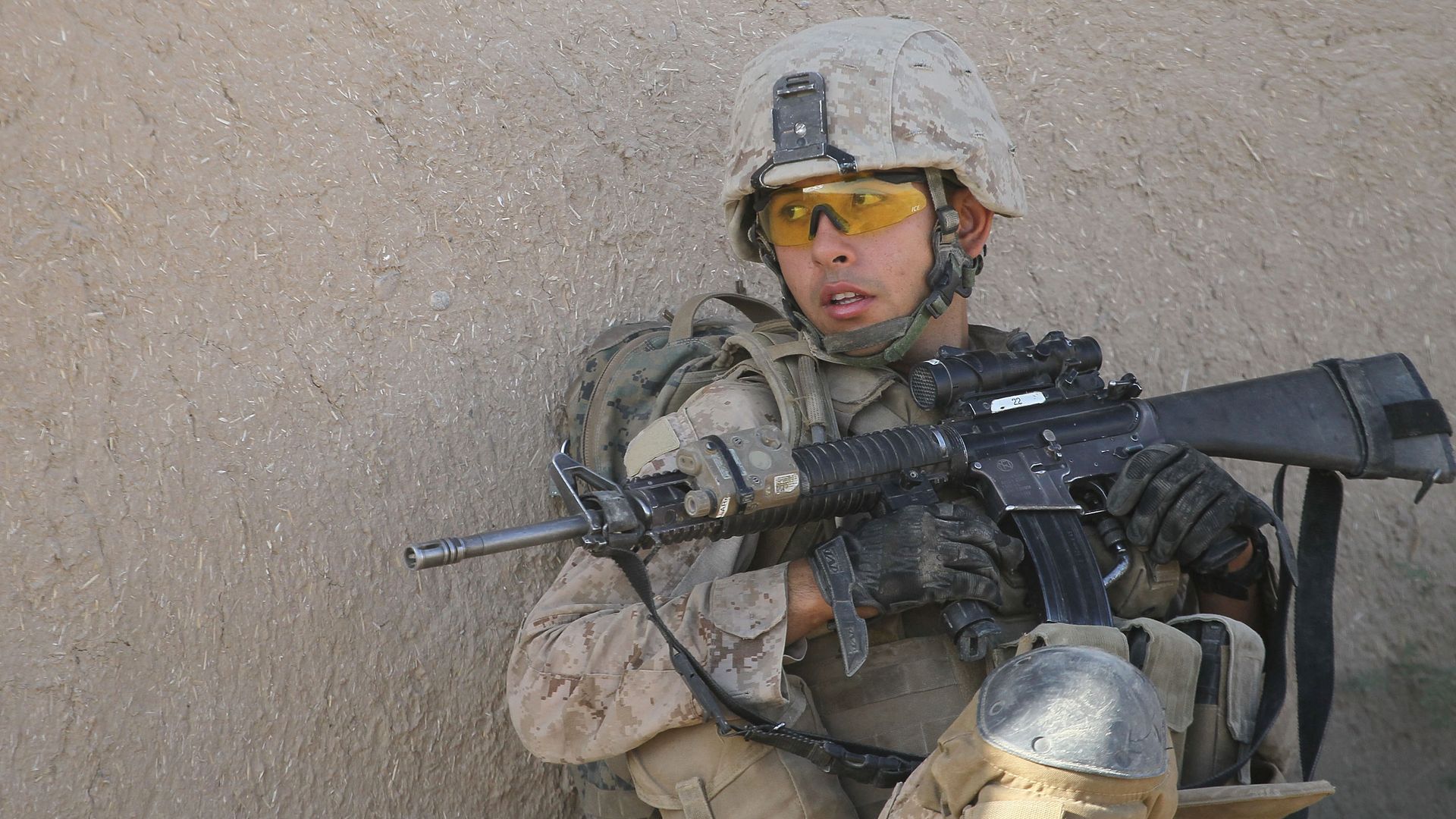 A soldier in camo uniform and helmet with yellow goggles holds a rifle, crouching a wall, appearing alert and ready.