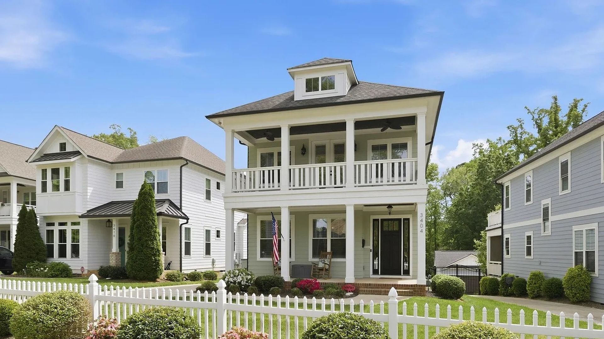 Two-story white house with a covered front porch and second-floor balcony, black front door, American flag, white picket fence, and manicured shrubs with pink flowers; blue sky above.