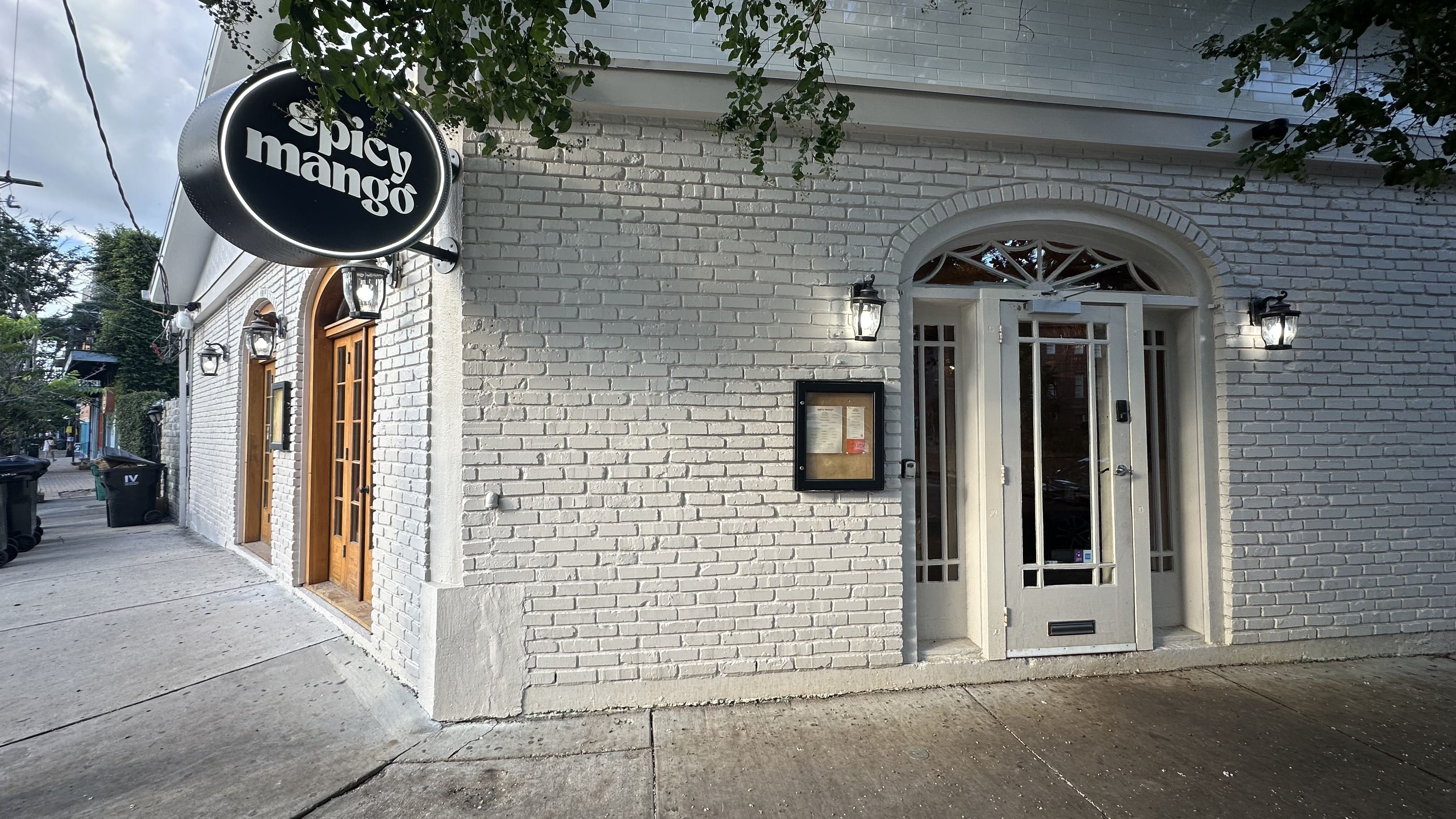 A white brick building has windows painted in orange. A sign hanging outside says Spicy Mango.
