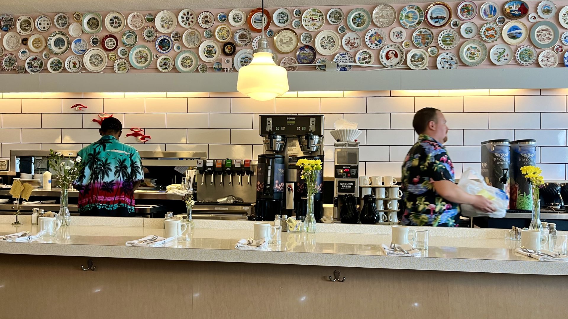 A breakfast counter with place settings, facing drink and coffee machines, a wall decorated with plates and two servers making runs in Hawaiian-print shirts.