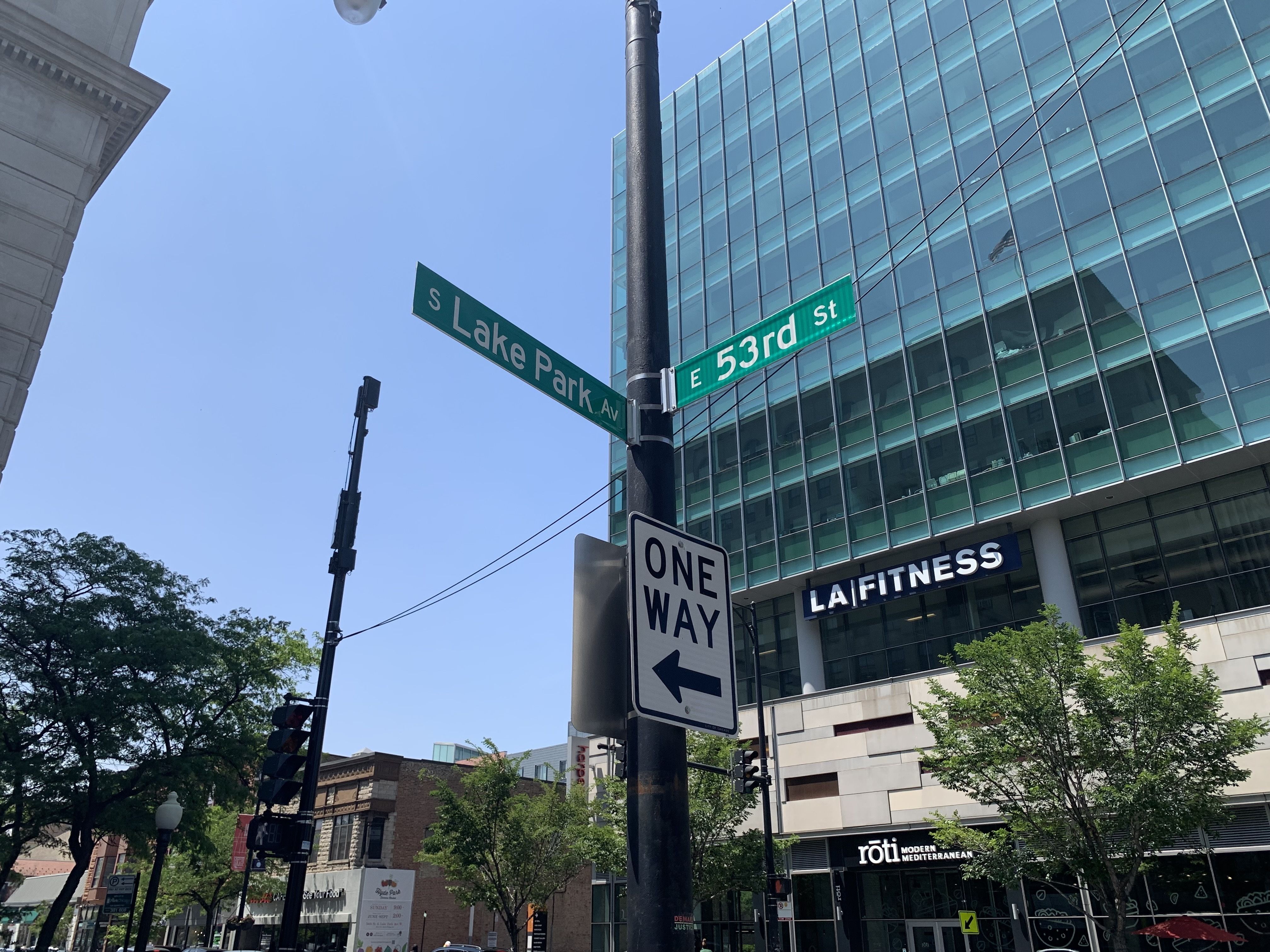 Street intersection at S Lake Park Ave and E 53rd St with one-way sign pointing left. Modern glass building with LA Fitness and Roti Mediterranean below, trees and clear blue sky.