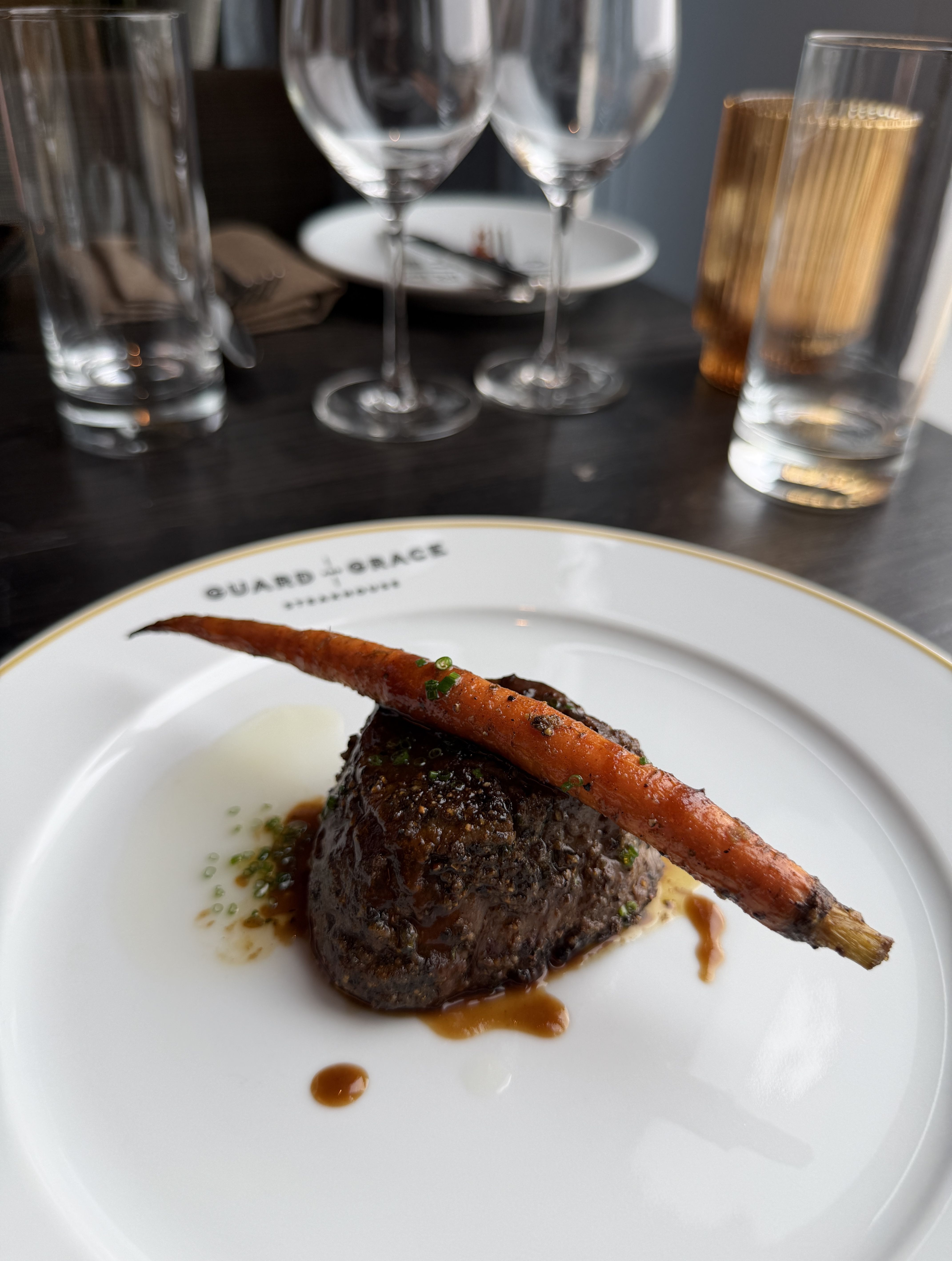 Plated beef steak with a long carrot garnish, drizzled with sauce and herbs, on a white plate; blurred background shows wine glasses and a gold-toned candle holder.