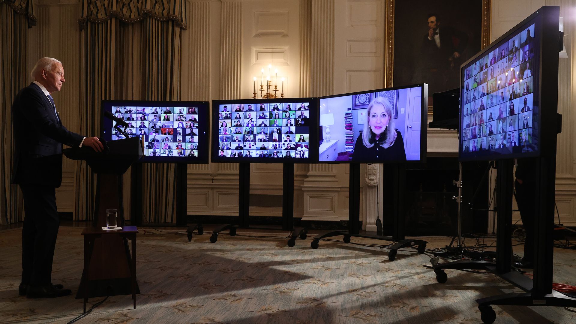 President Biden is seen standing before three TV screens as he conducts a virtual swearing-in for some of his staff.