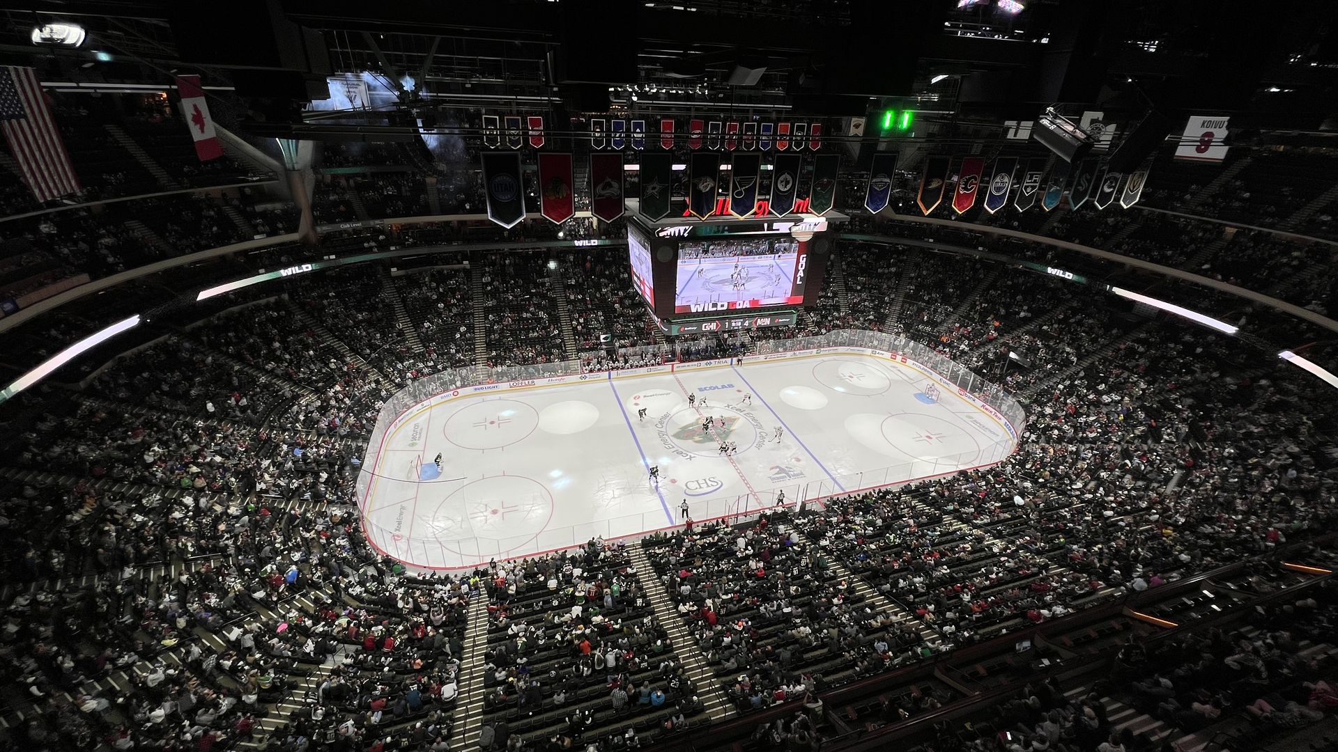 An NHL hockey arena seen from high overhead during a game, lit with bright spotlights as a crowd cheers in the seats below