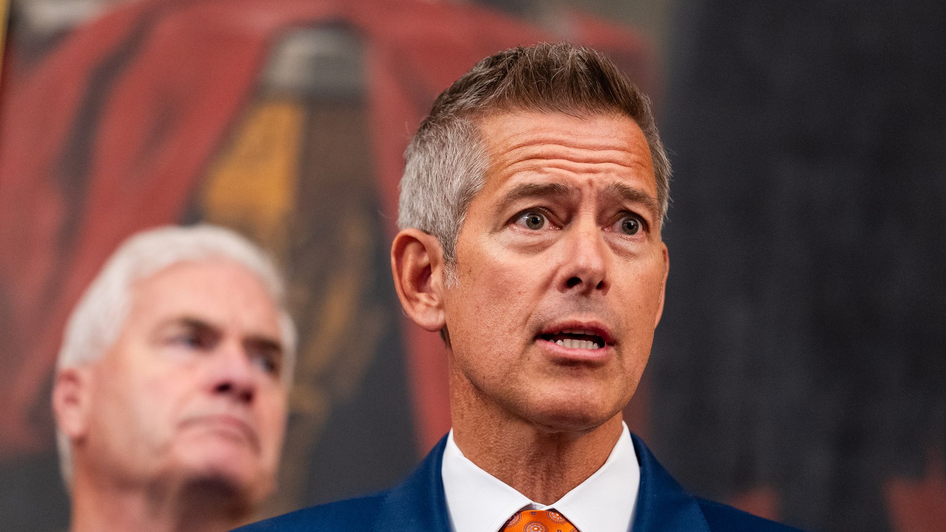 Duffy wearing a blue suit, white shirt and orange tie